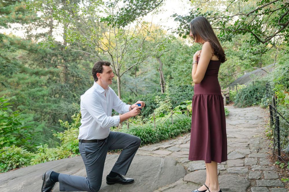 Man proposing on one knee to woman in burgundy dress on a stone path in a green park.