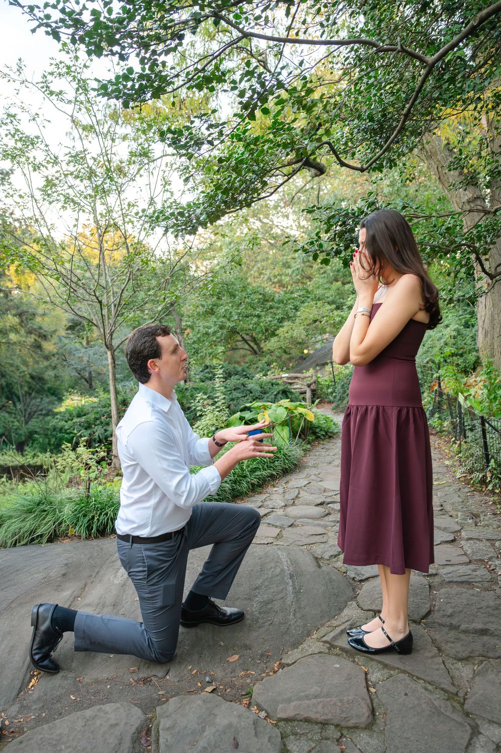 Man kneeling on one knee proposing to surprised woman in burgundy dress on stone path in lush garden.