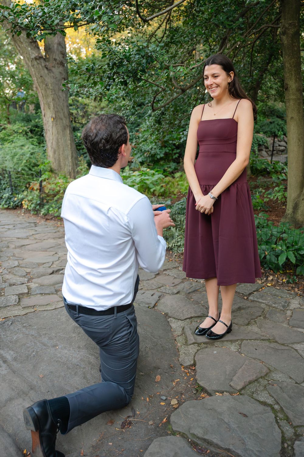Man kneeling on one knee proposing to woman in burgundy dress in garden.