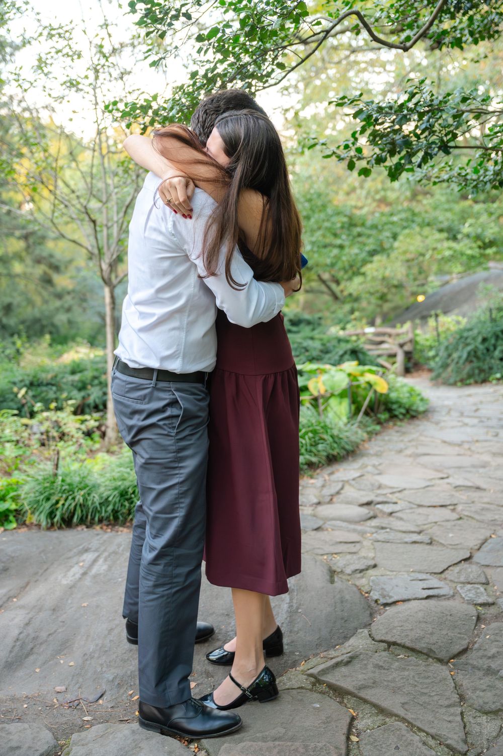 Couple embracing and hugging on a stone garden path surrounded by lush green trees and plants.
