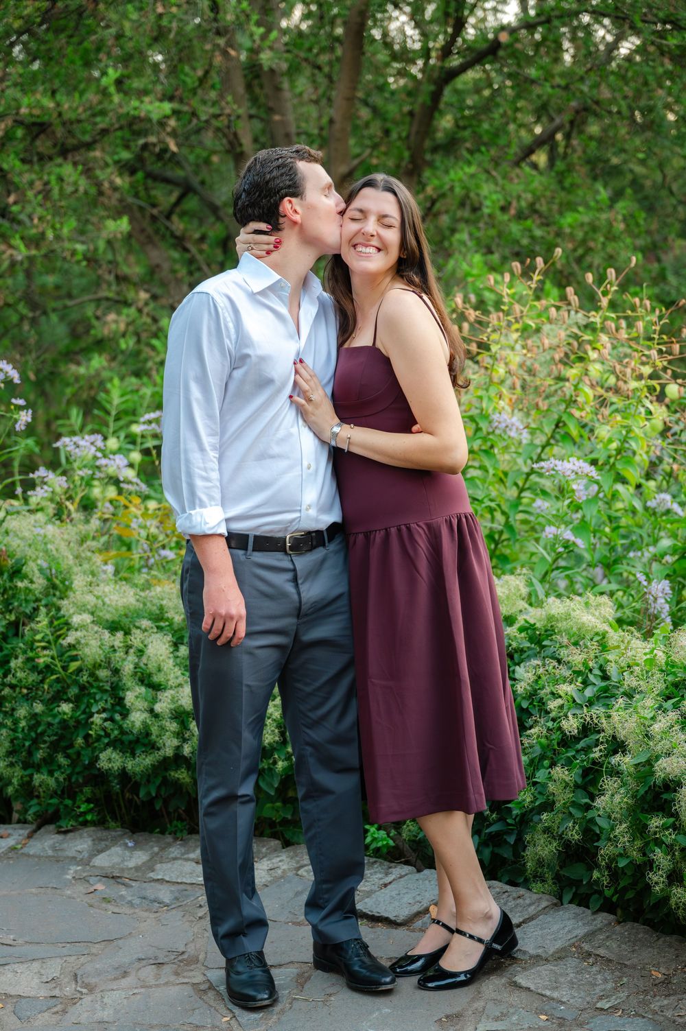 Couple sharing a romantic kiss in a lush garden, man in white shirt and woman in burgundy dress smiling.