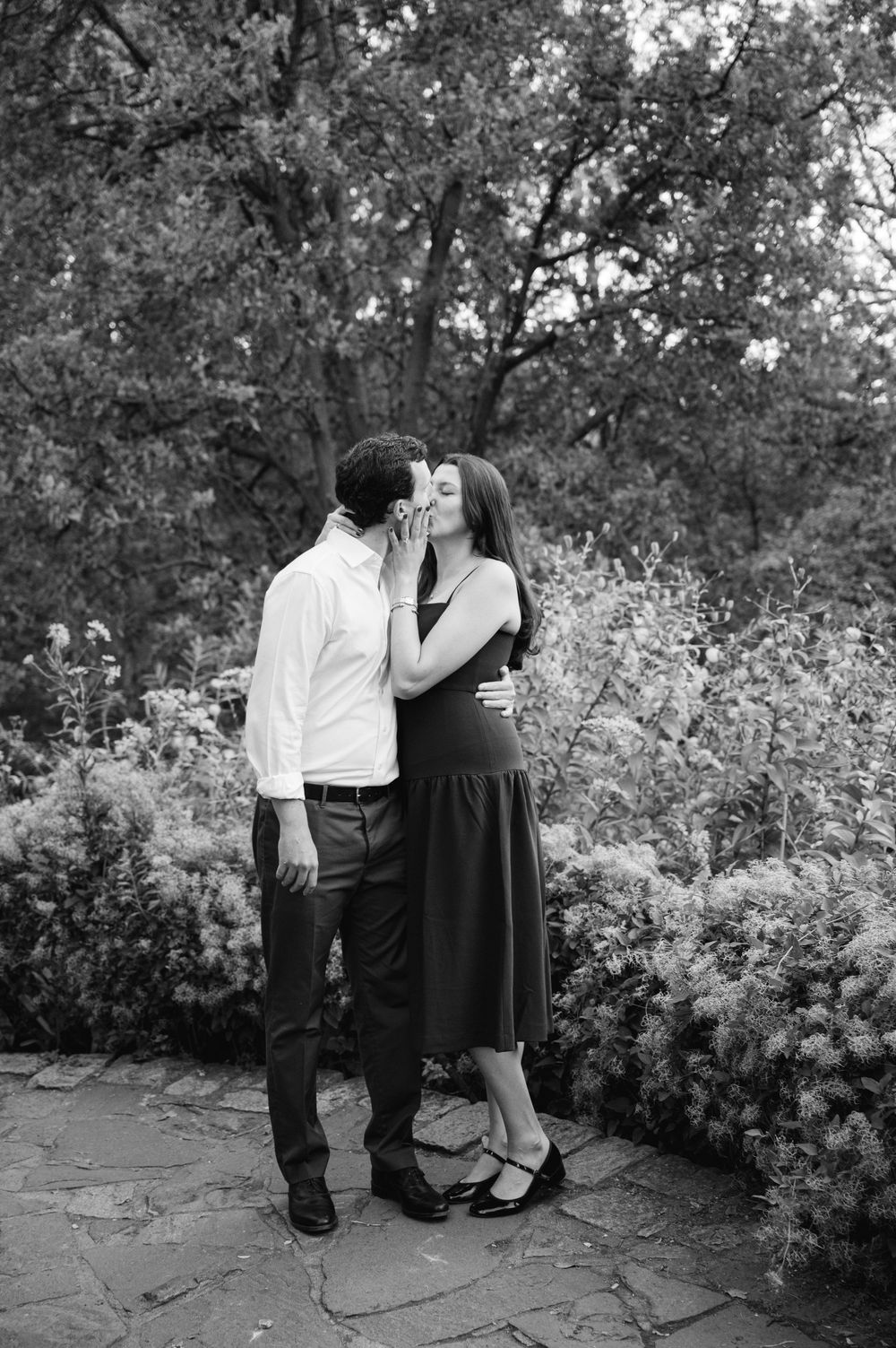 Black and white photo of couple sharing a tender kiss in a lush garden setting.
