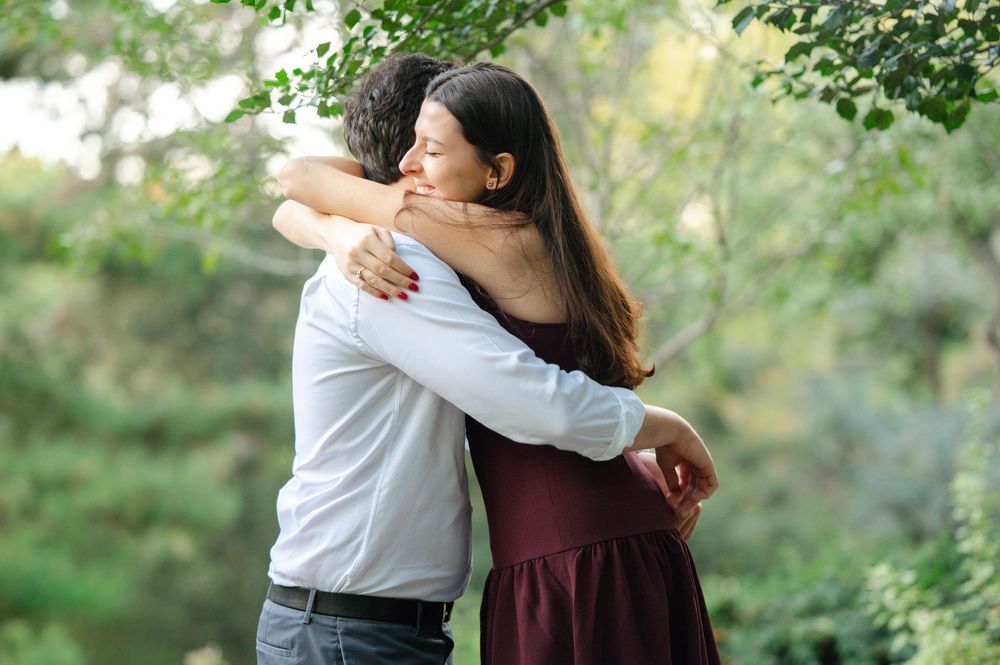 Couple embracing in a warm hug surrounded by lush green trees outdoors.