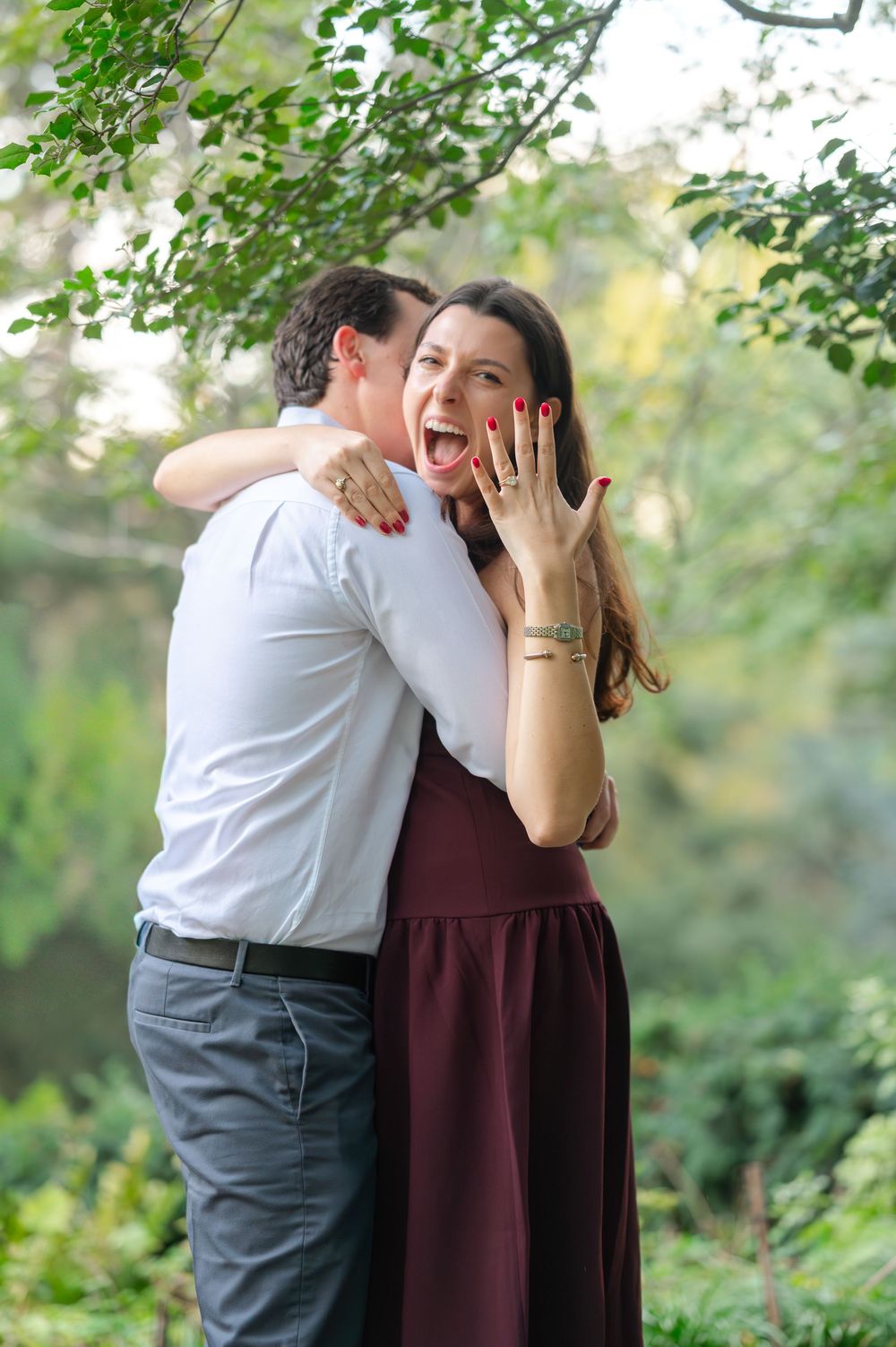 Excited woman shows off engagement ring while man embraces her in a green outdoor setting.