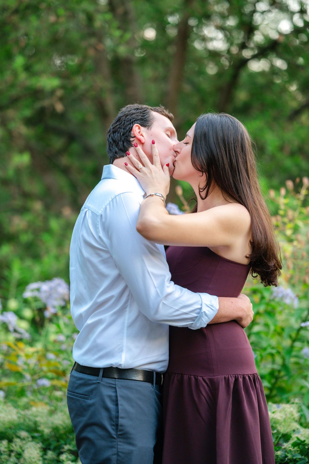 Couple sharing a romantic kiss in a lush garden, woman in burgundy dress embracing man in white shirt.