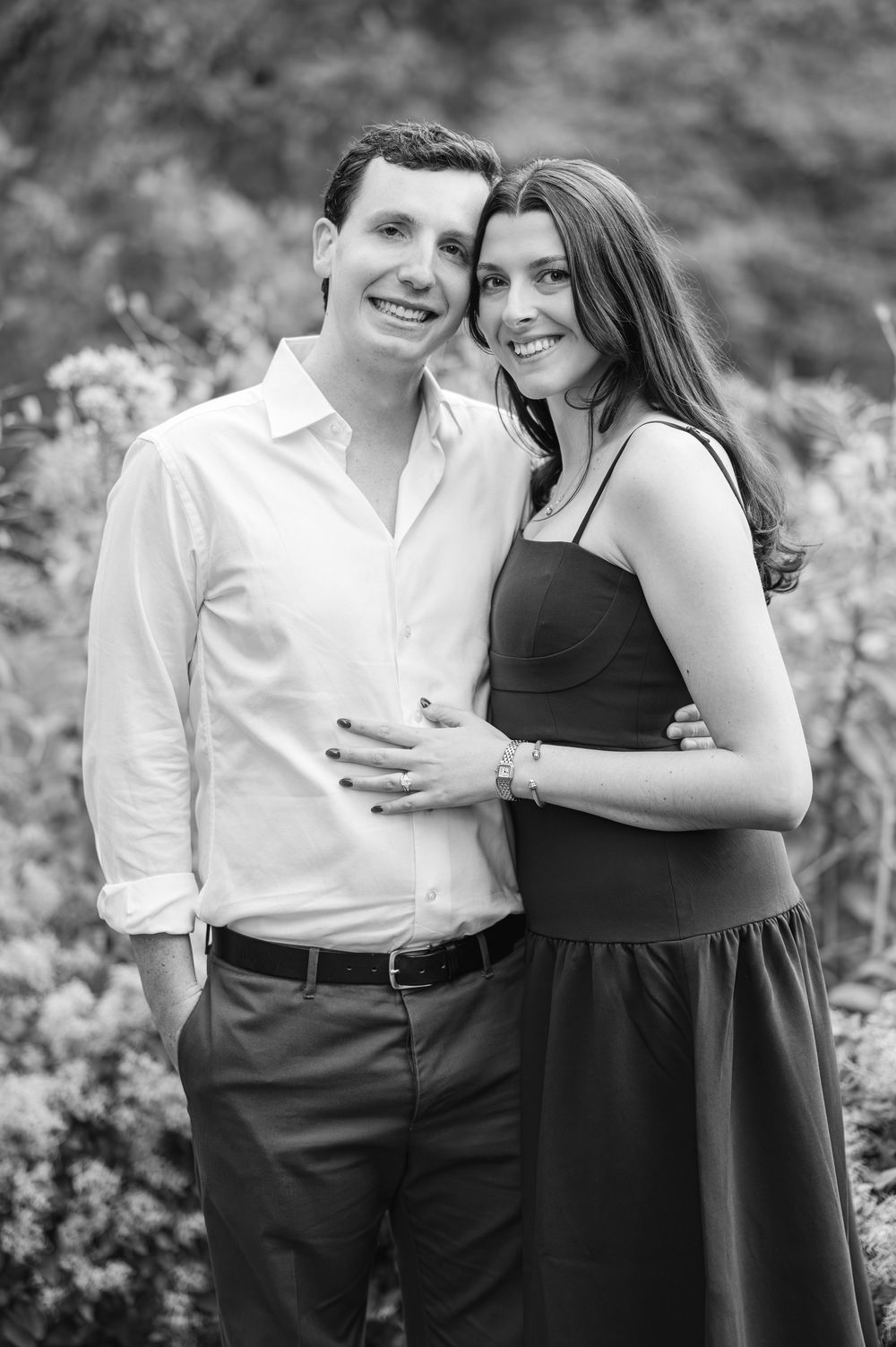 Black and white portrait of a smiling couple embracing outdoors among blooming flowers.
