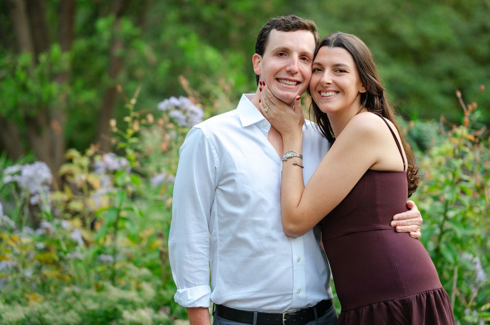 Smiling couple embracing outdoors in a lush garden, woman in burgundy dress rests hand on mans cheek.