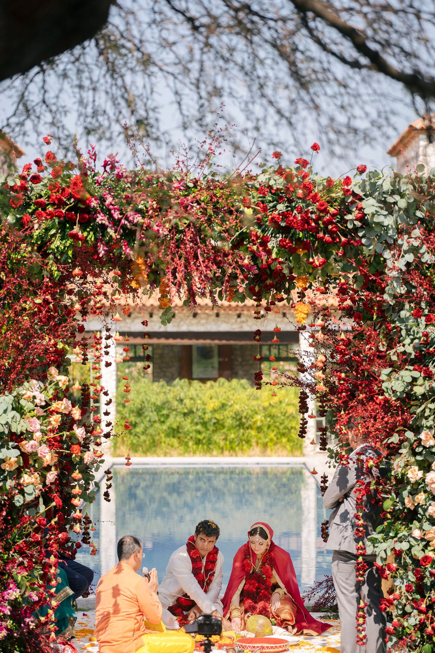 Indian wedding ceremony under floral arch with red blooms, bride in red saree and groom seated with priest outdoors.