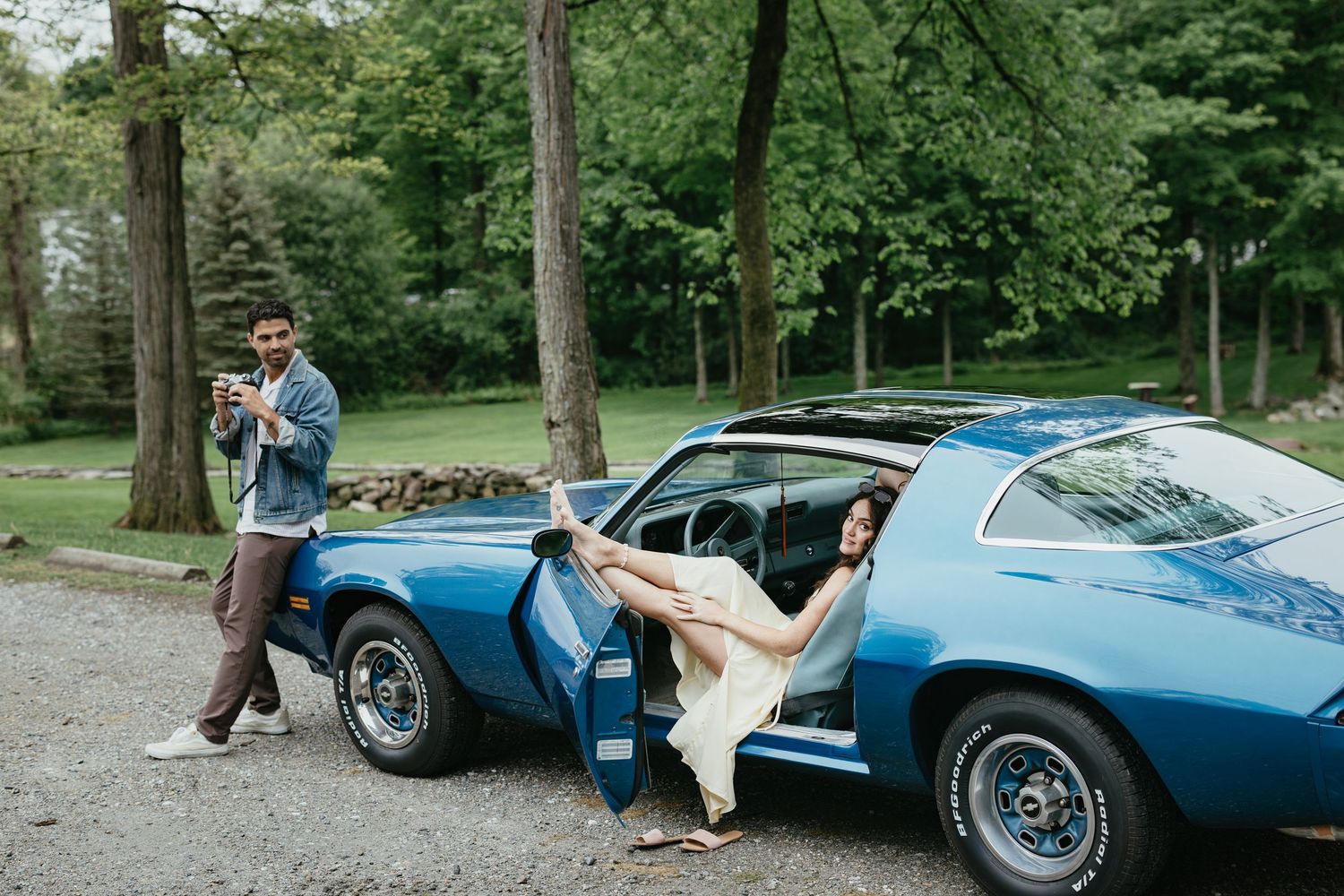 Couple posing with a classic blue muscle car outdoors, man photographing woman sitting in open car door.