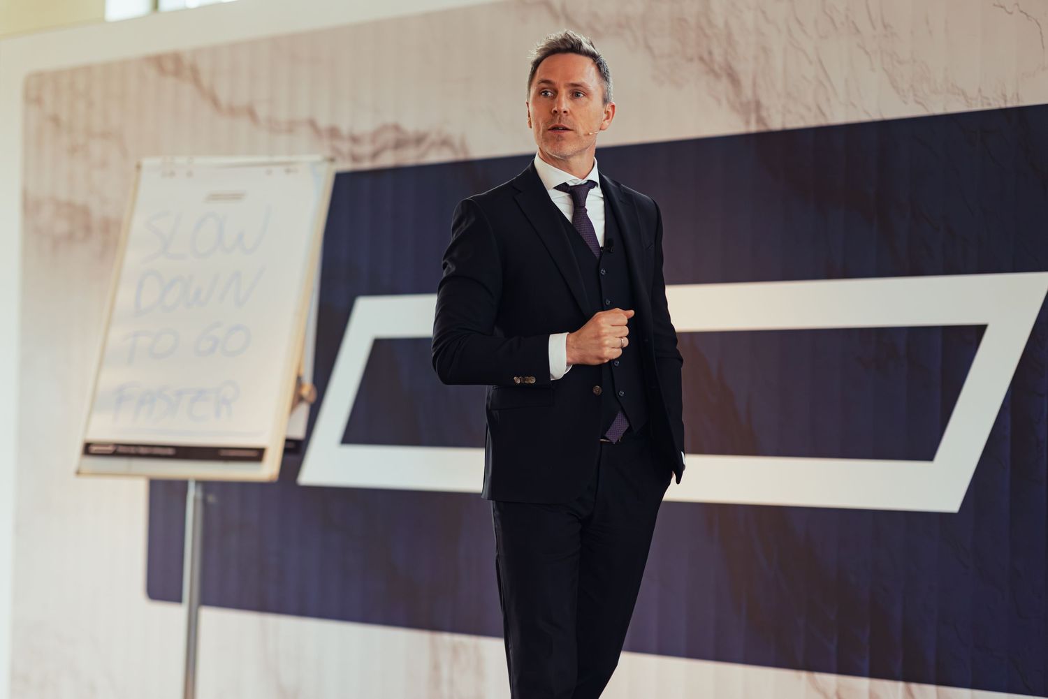Man in navy three-piece suit smiling during a professional business presentation in a modern conference room.
