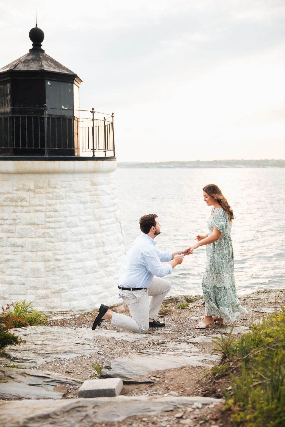 Man proposing on one knee to woman in green dress by waterfront lighthouse at sunset.  , Castle Hill.  Engagement photography Newport RI