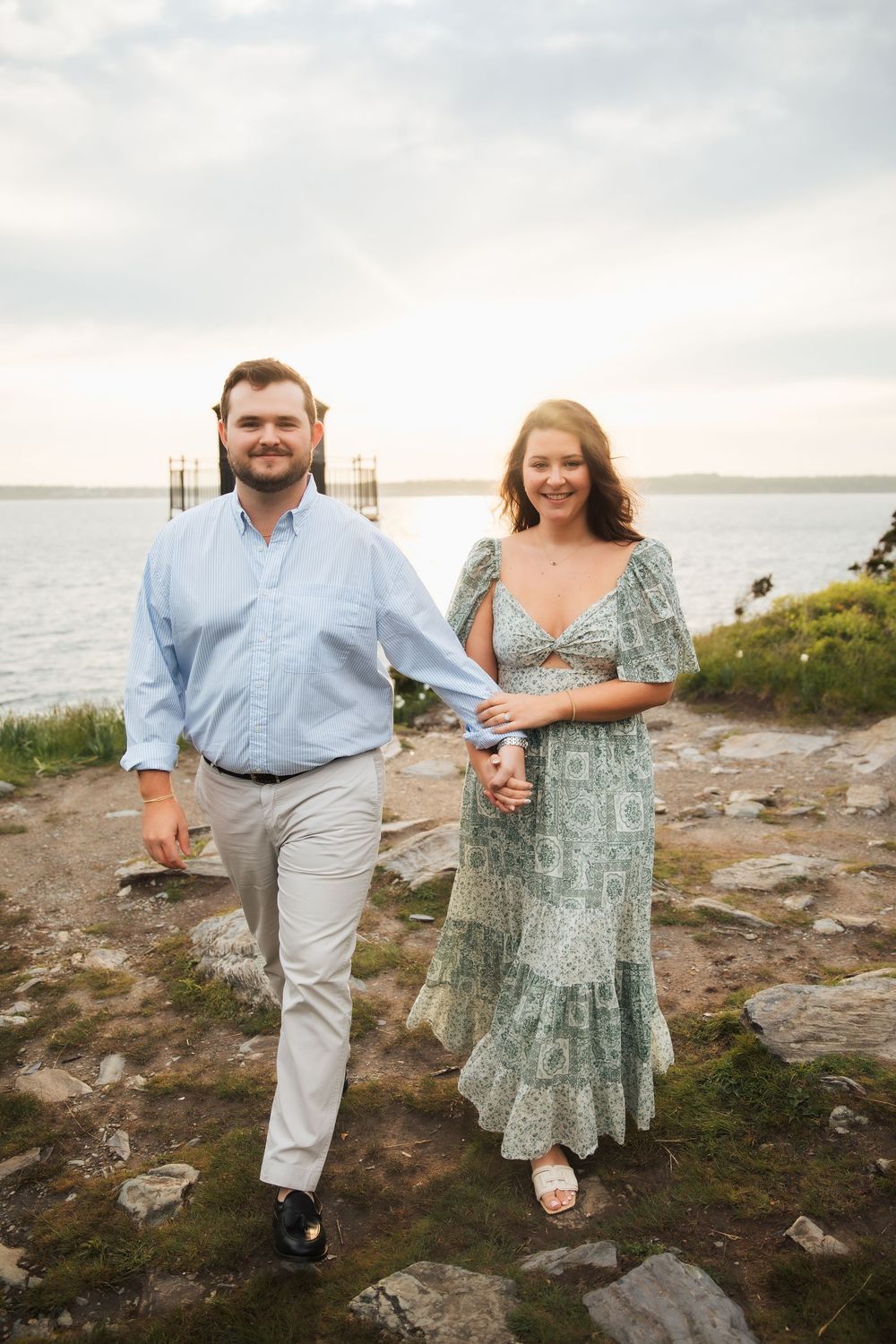 Couple holding hands walking along rocky coastal path at sunset, woman in floral green dress. Engagement photography Newport RI