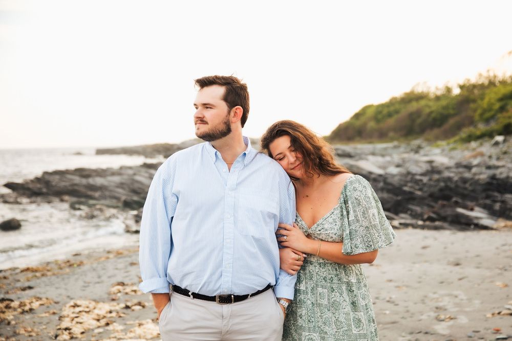 Couple embracing on rocky beach at sunset, woman in floral dress leaning on man in light blue shirt. Engagement photography Newport RI