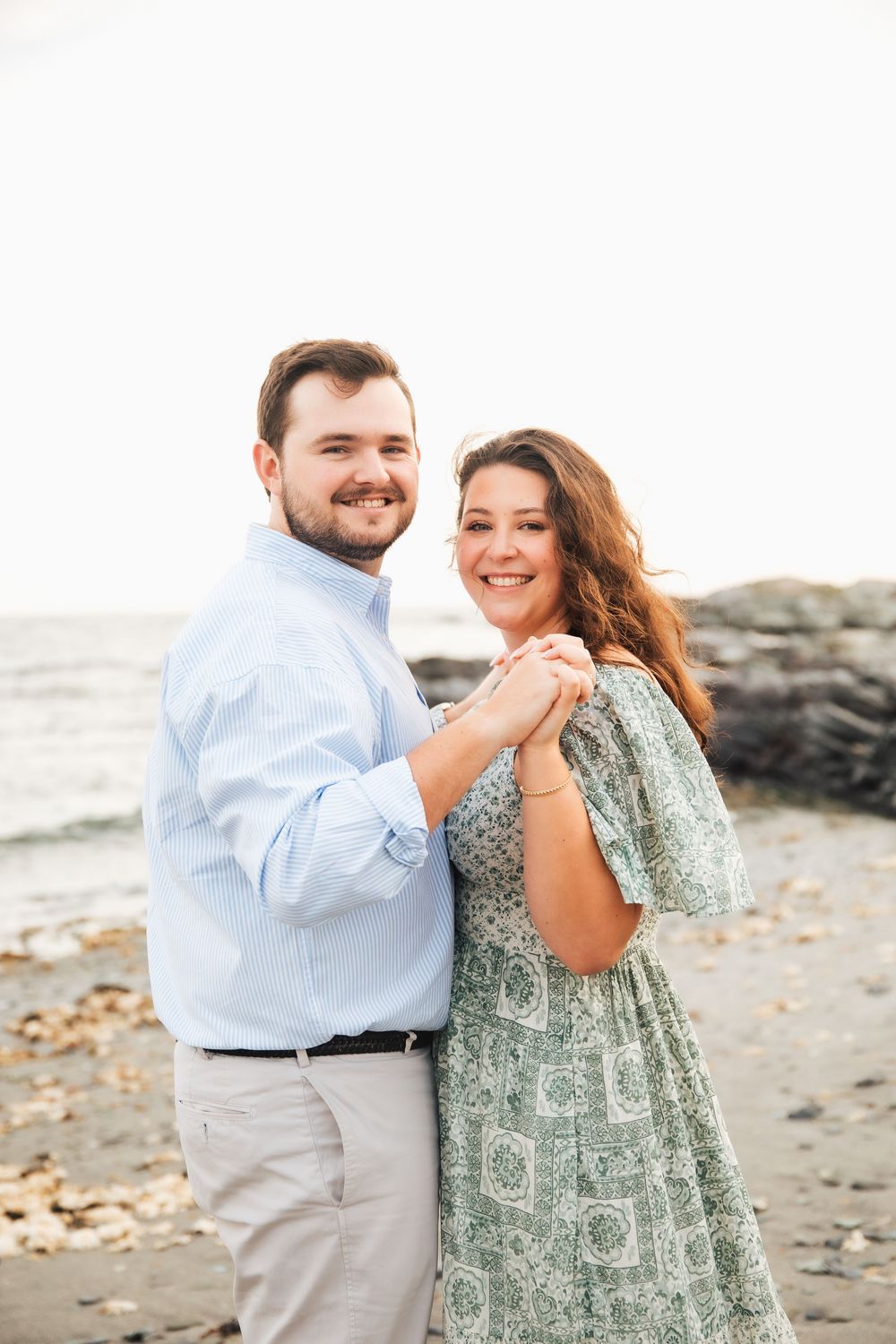 Happy couple holding hands and smiling on a rocky beach, man in blue shirt and woman in floral dress. Engagement photography Newport RI