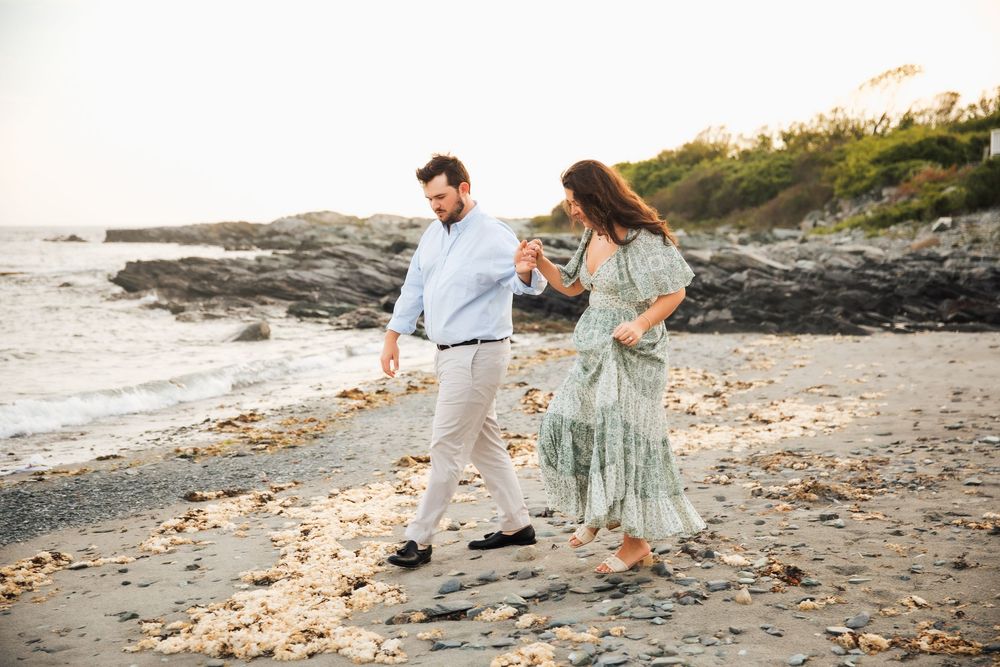 Couple walking hand in hand on a rocky beach at sunset, woman in green floral dress, man in blue shirt. Engagement photography Newport RI