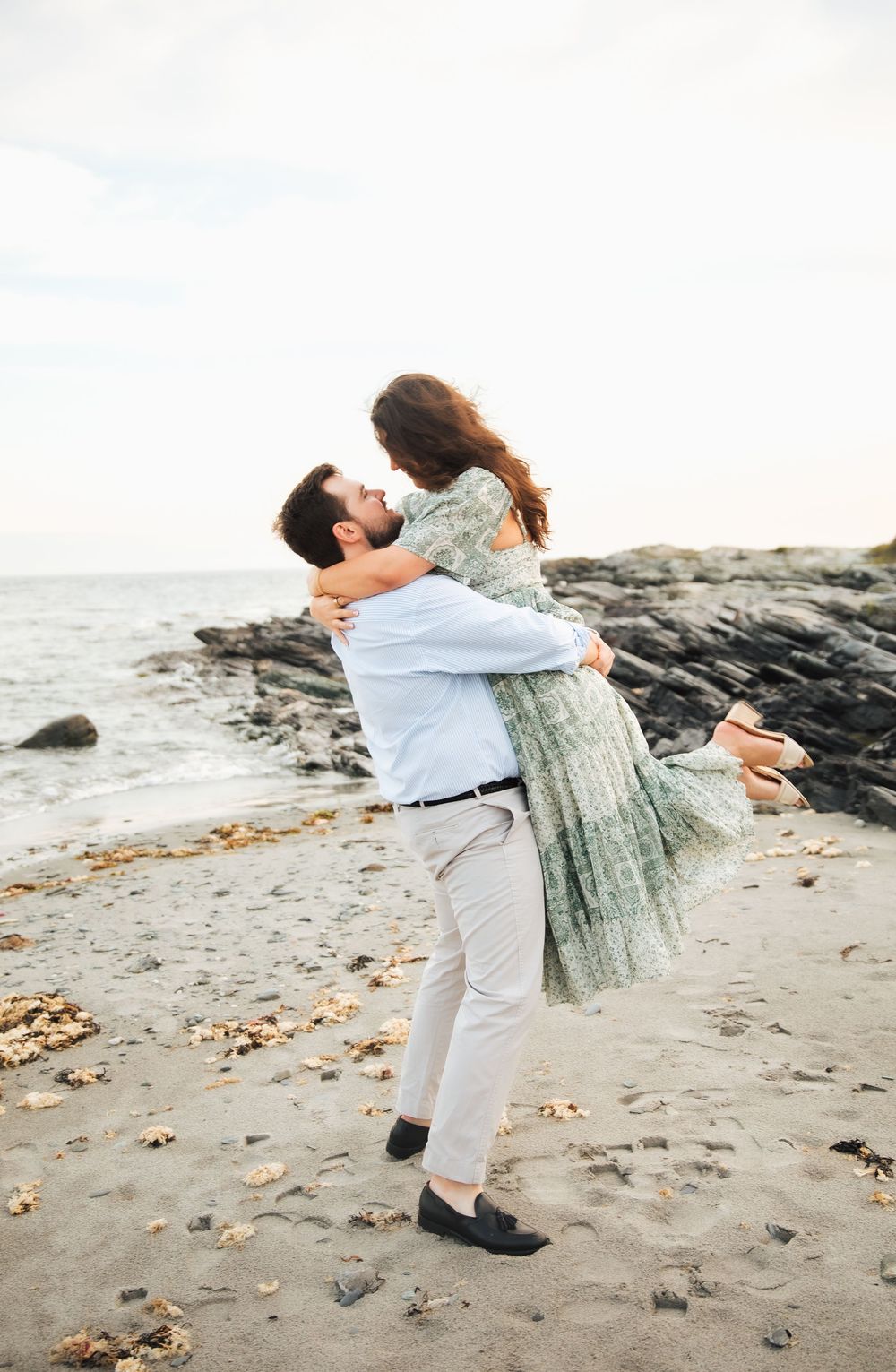 A man lifts a woman in a green dress on a rocky beach, sharing a romantic embrace by the ocean. Engagement photography Newport RI