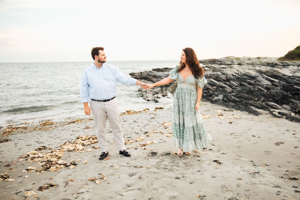Couple holding hands on a rocky beach at sunset, man in blue shirt and woman in floral green dress. Engagement photography Newport RI