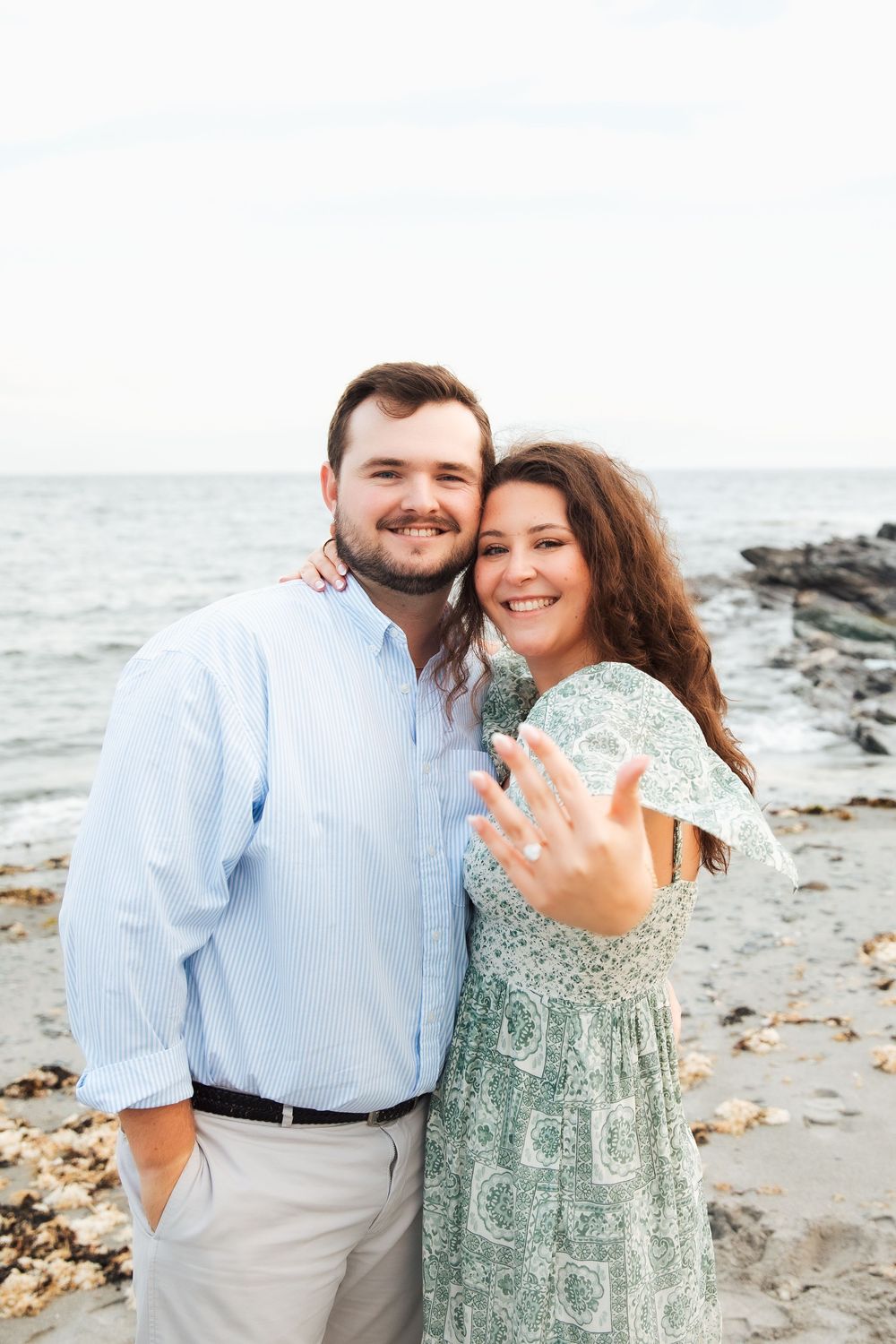 Happy couple at beach showing engagement ring, woman in floral dress, man in blue shirt, ocean backdrop. Engagement photography Newport RI