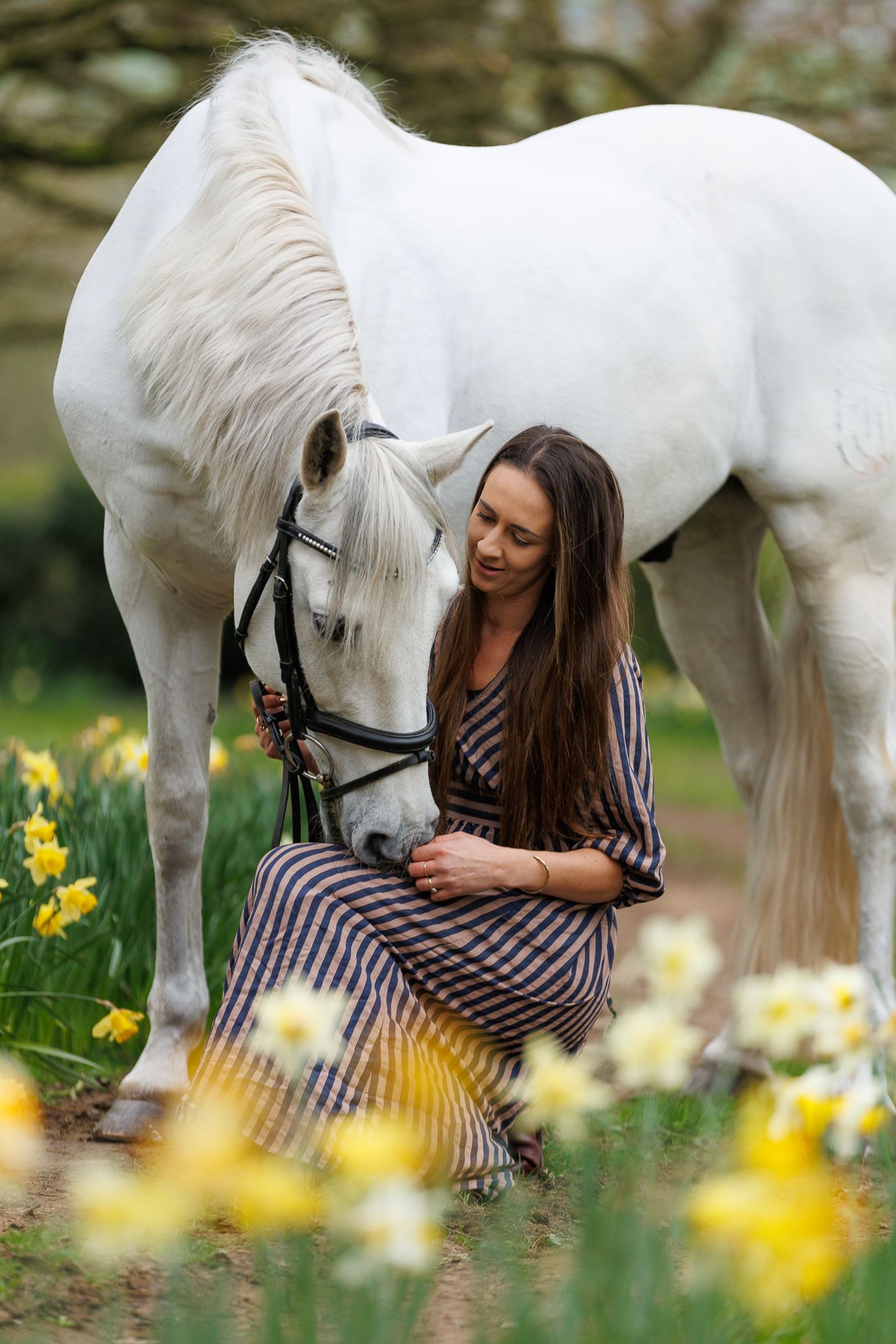 Black and white photo of a smiling girl in white outfit walking with a white pony on a tree-lined path.