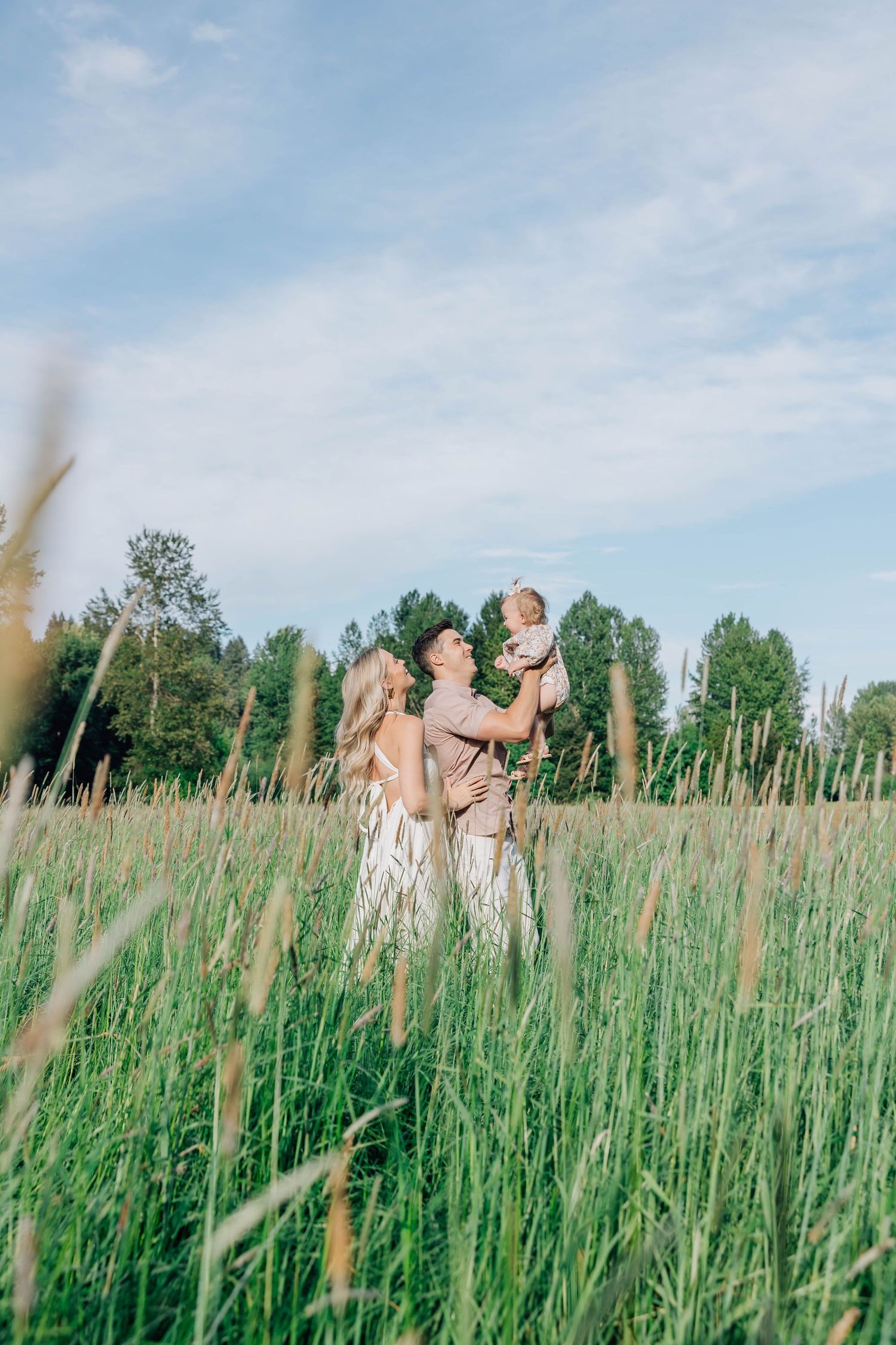 A couple stands in a lush green field, joyfully lifting their baby up toward the blue sky.
