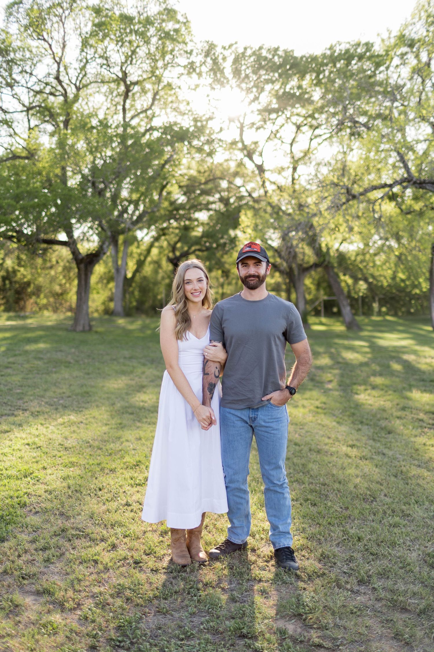 Couple posing outdoors in a sunny park, woman in white dress and man in gray shirt and jeans, surrounded by trees at Commons Ford Ranch.