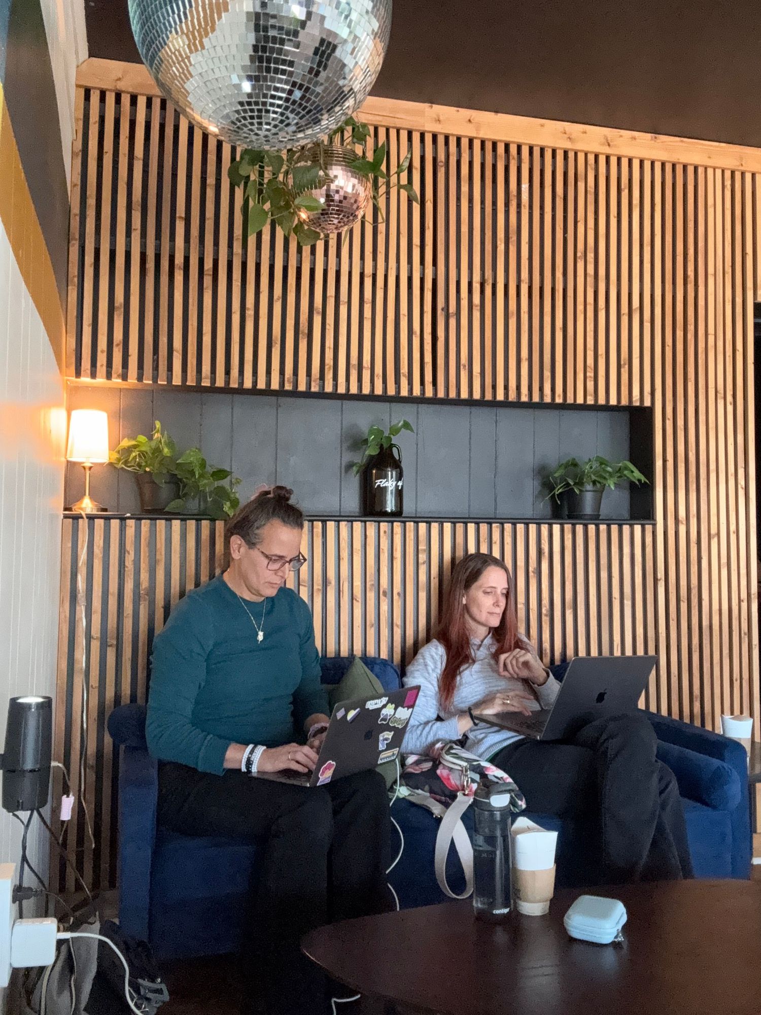 Two women sitting on blue velvet sofas working on laptops in a modern cafe with wooden slat walls and a disco ball.