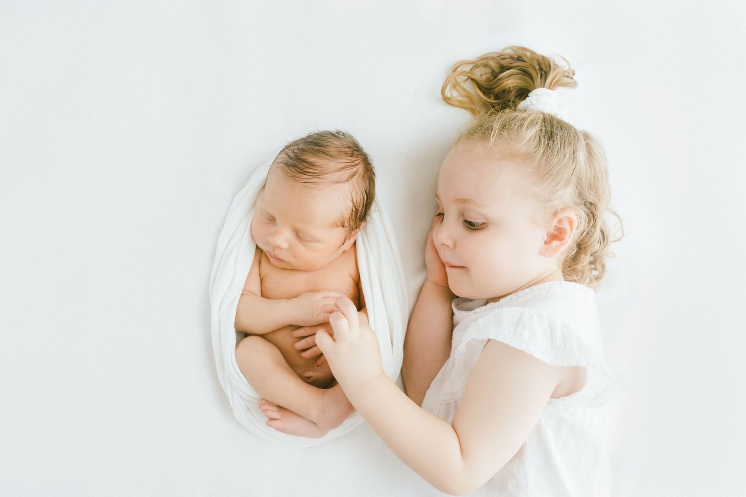 Toddler girl in white dress gazes lovingly at sleeping newborn baby wrapped in white swaddle.