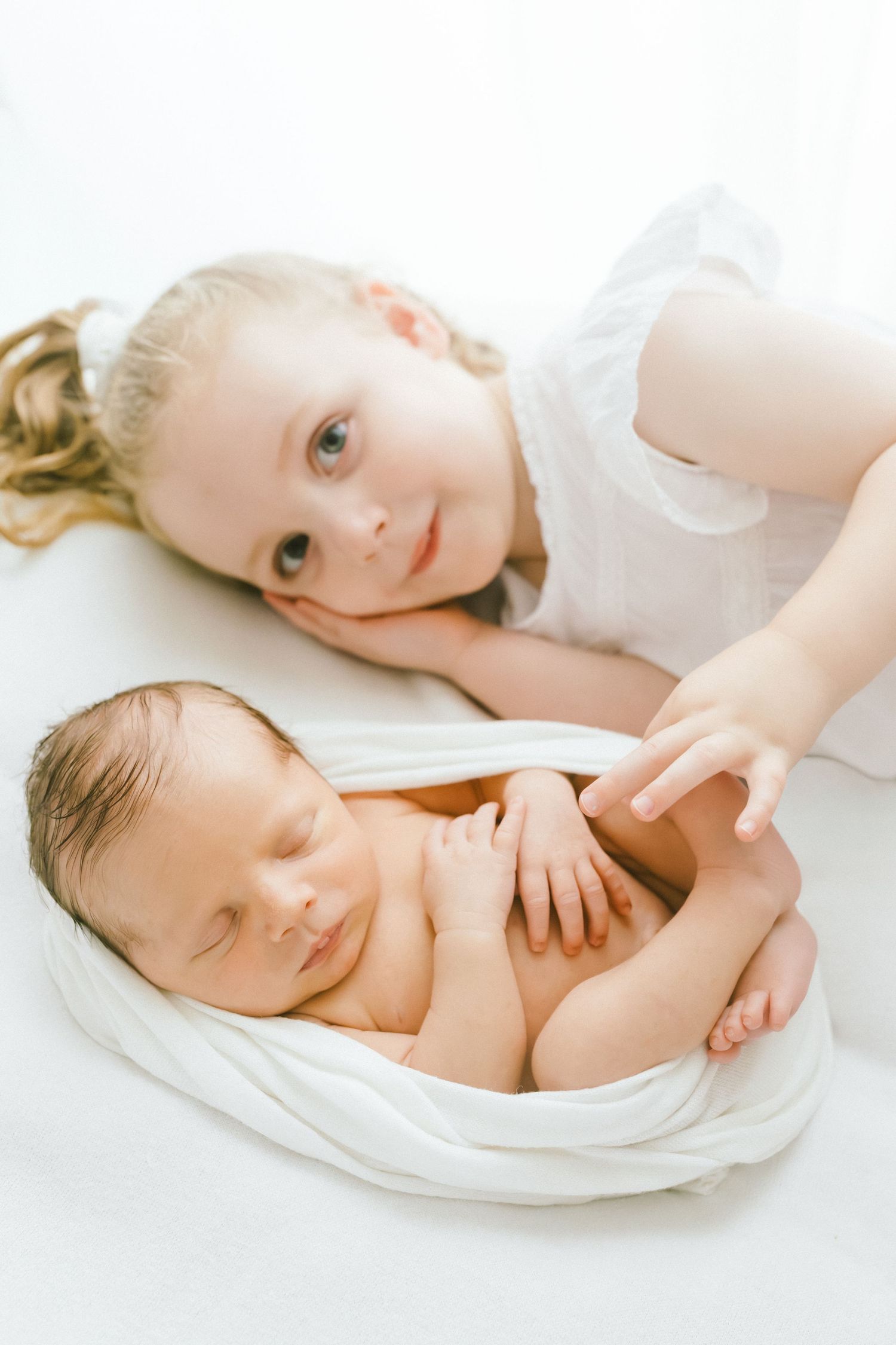 Young girl in white dress tenderly touches sleeping newborn baby wrapped in white cloth on white background.