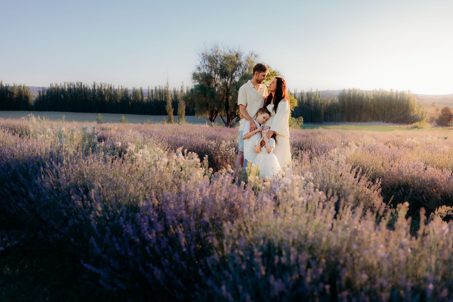 Pregnant woman in cowboy hat and jeans leans against tree in golden sunset field for maternity photo.