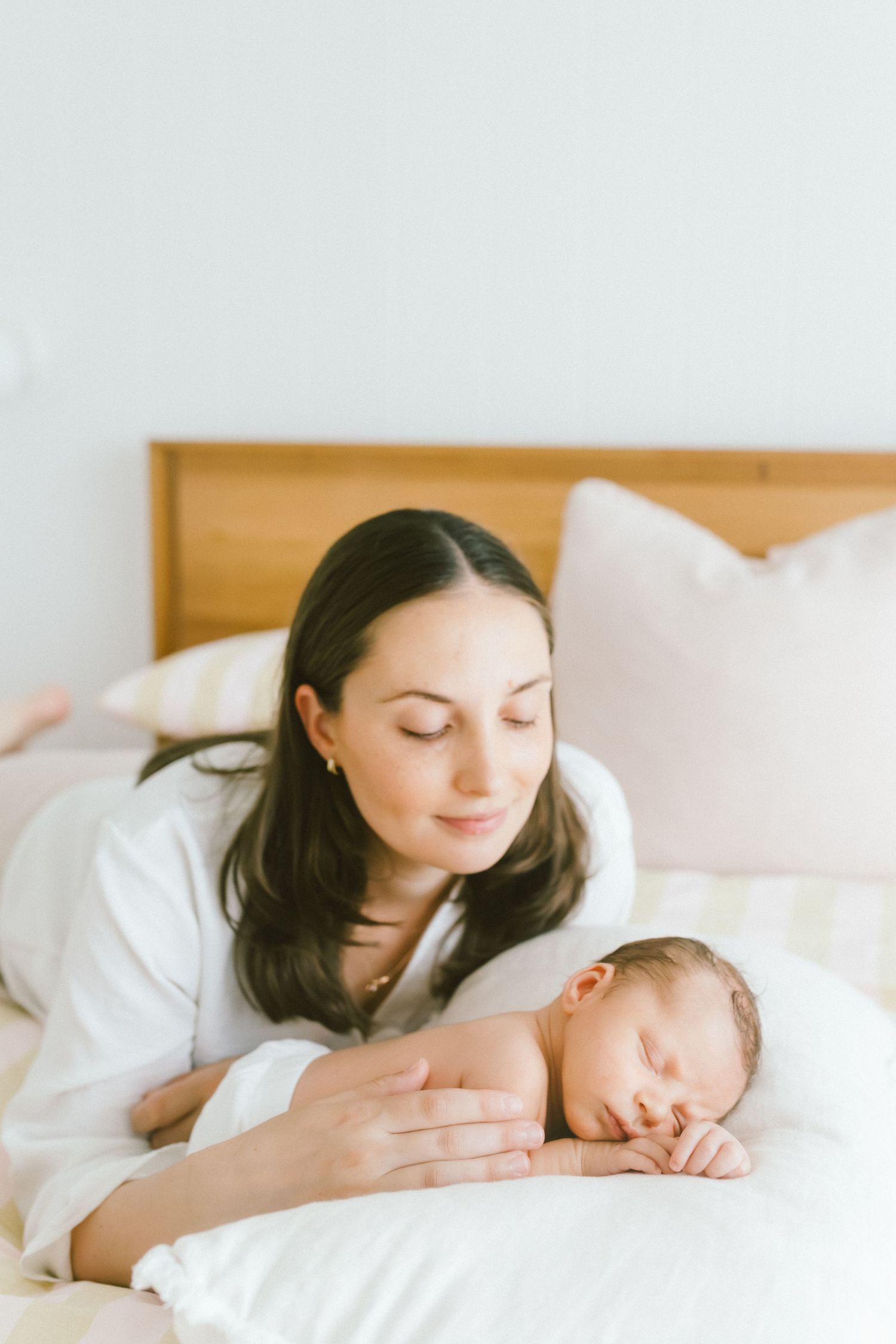 A mother with dark hair gazes lovingly at her sleeping newborn baby on a white bed.