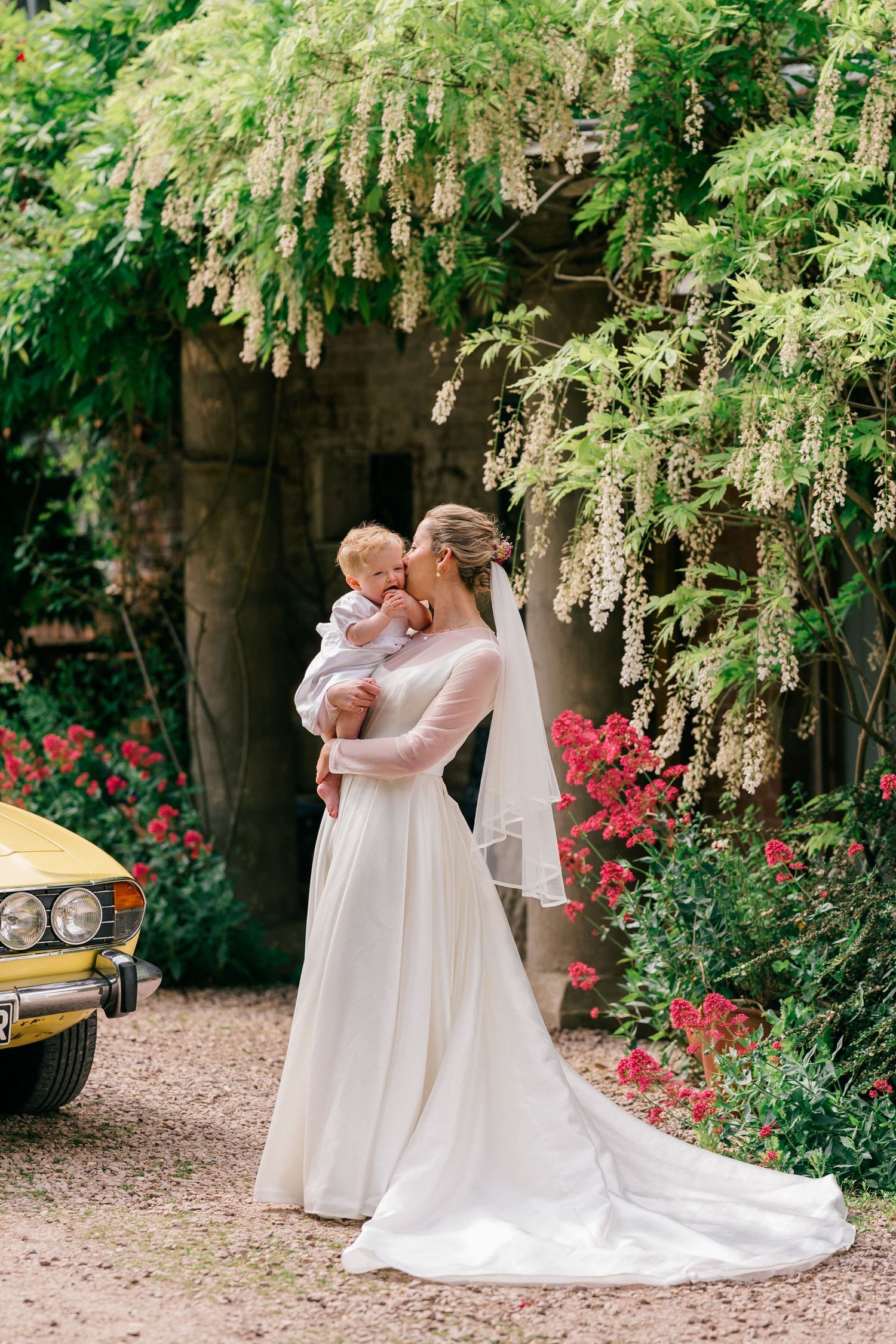 Bride in white gown holding baby amid lush garden with wisteria and red flowers beside vintage yellow car.