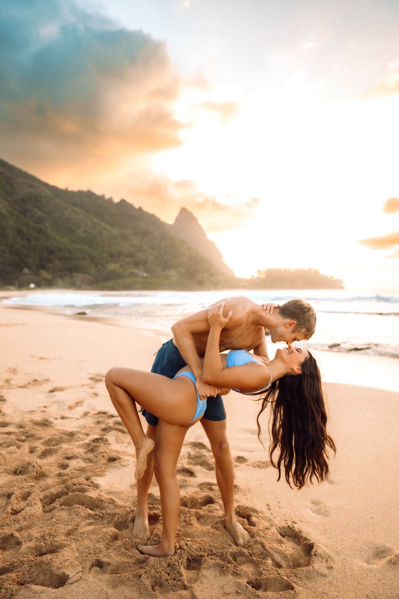Romantic couple kissing on a tropical beach at golden sunset with lush green mountains in the background.