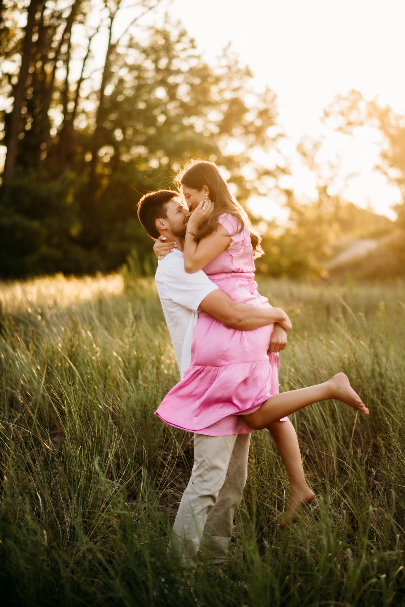 Couple sharing a romantic kiss in a sunlit field, man lifting woman in pink dress at golden hour.