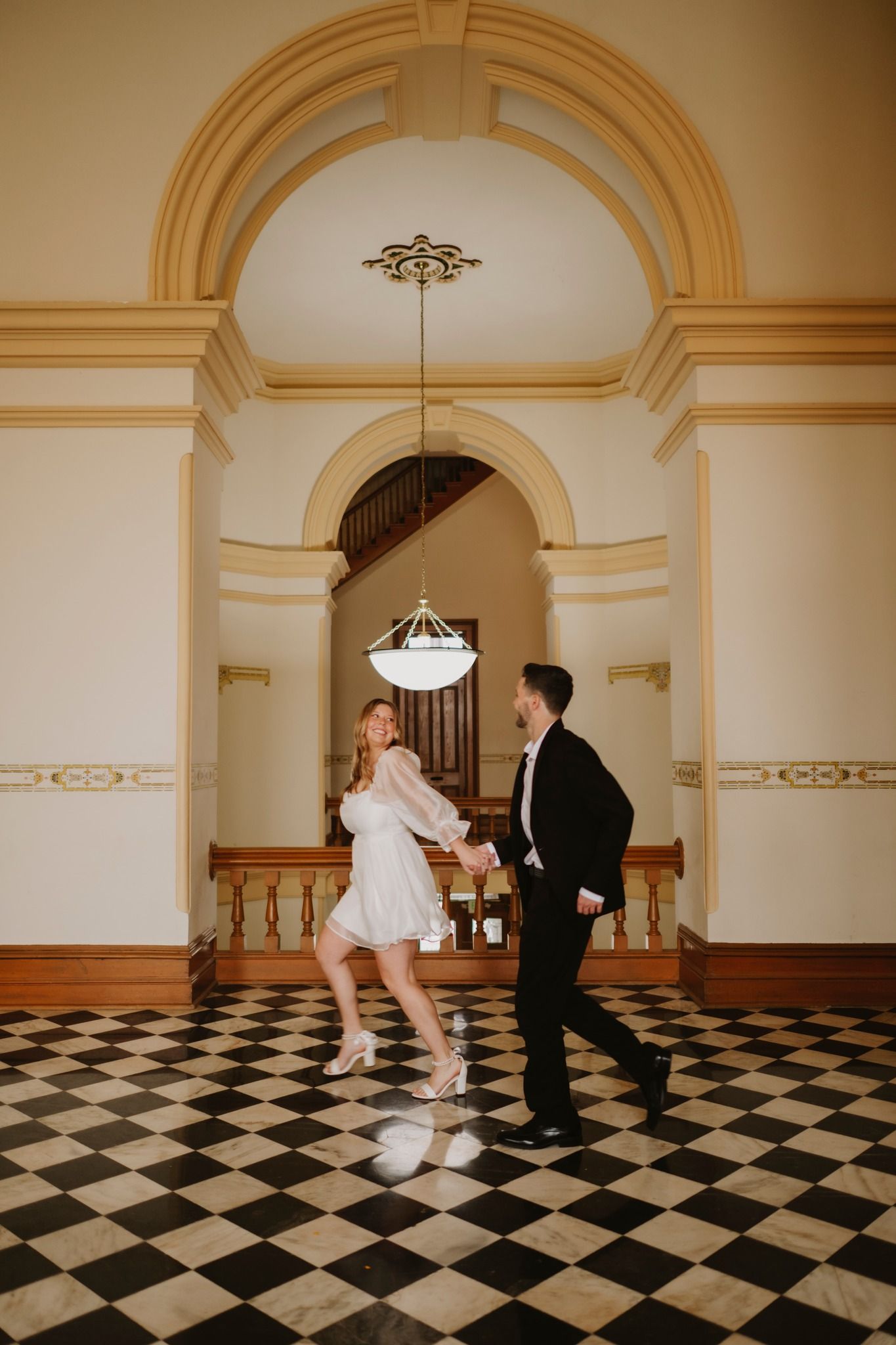 Couple dancing joyfully in elegant venue with checkered floors, arched doorways, and ornate moldings.