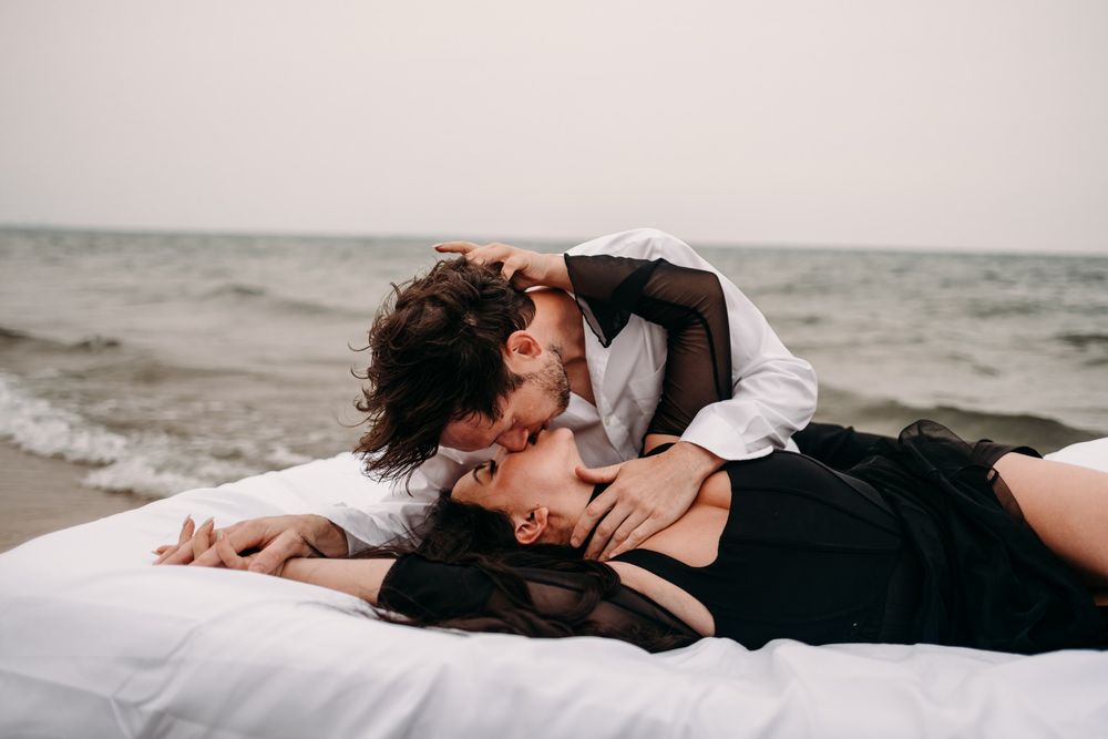 Romantic couple kissing on white bedding at beach, woman in black dress, man in white shirt, ocean backdrop.