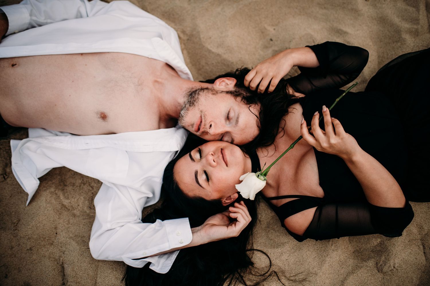 Couple lying on sand beach, woman in black dress holding white rose, man in open white shirt, romantic outdoor portrait.