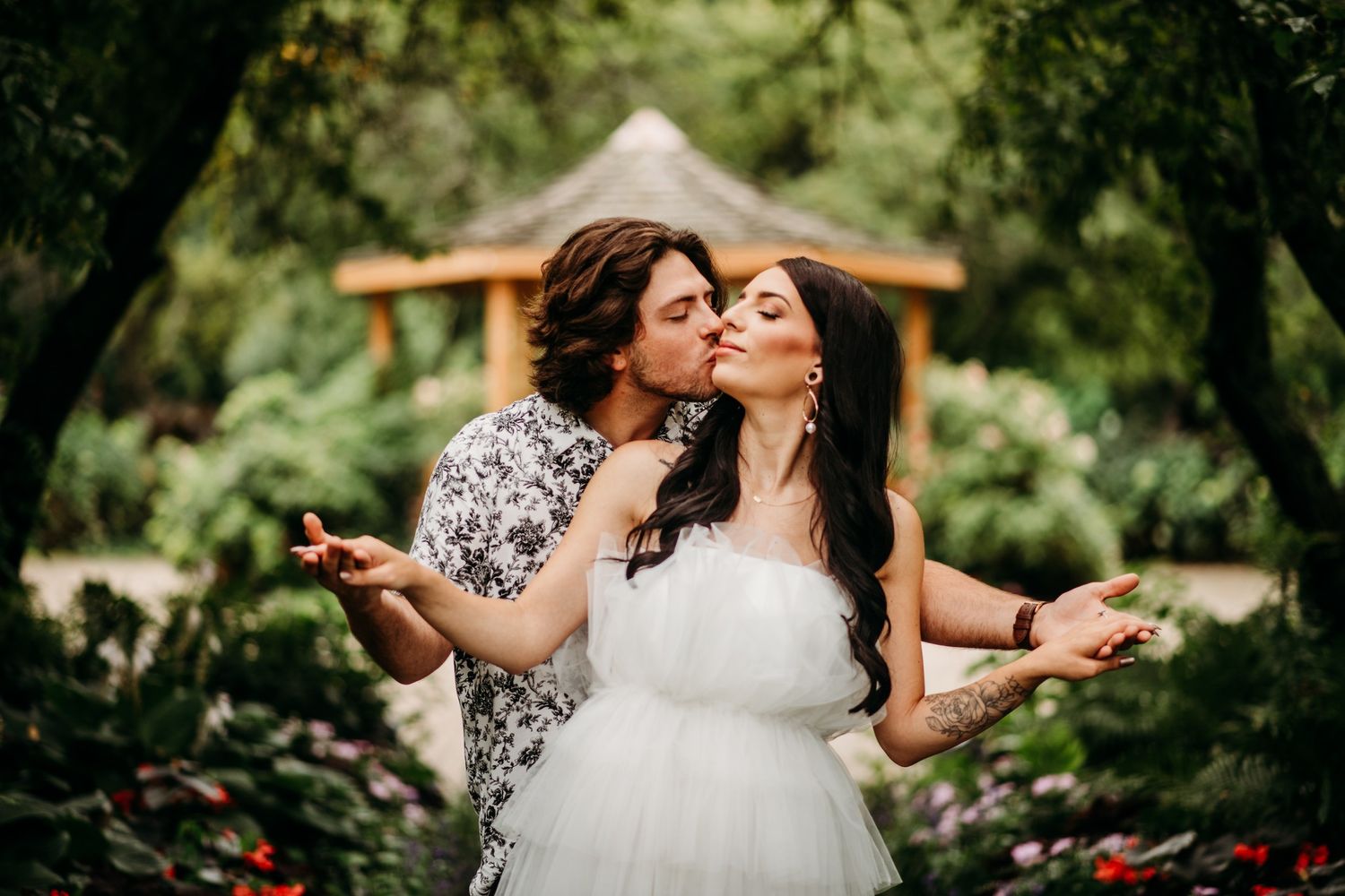 Couple sharing a romantic kiss in a lush garden, bride in white dress with floral tattoo, groom in floral shirt.
