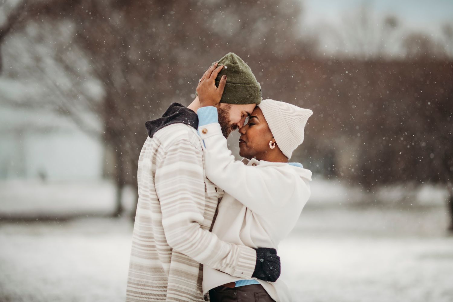 Romantic couple embracing in snowy winter park, wearing cozy hats and winter coats in snowfall.
