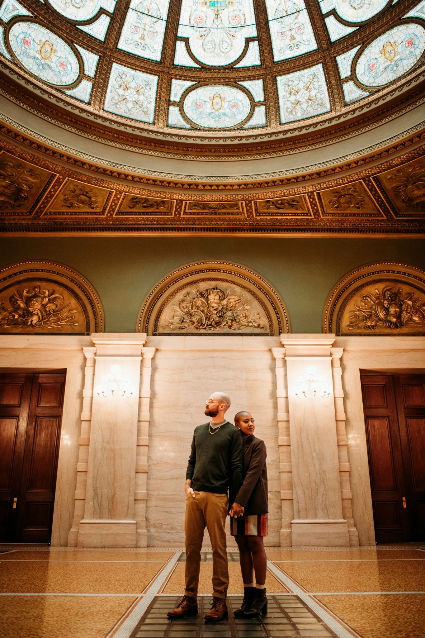 Couple holds hands under ornate domed ceiling with stained glass in elegant marble rotunda.