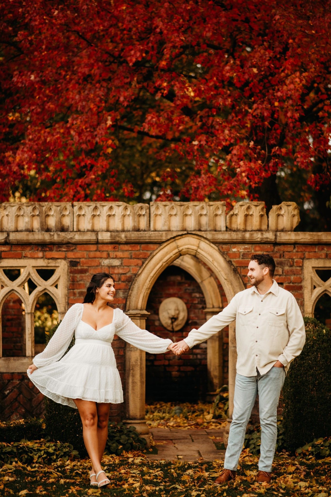 Couple holds hands in front of Gothic stone fountain surrounded by vibrant red autumn foliage.