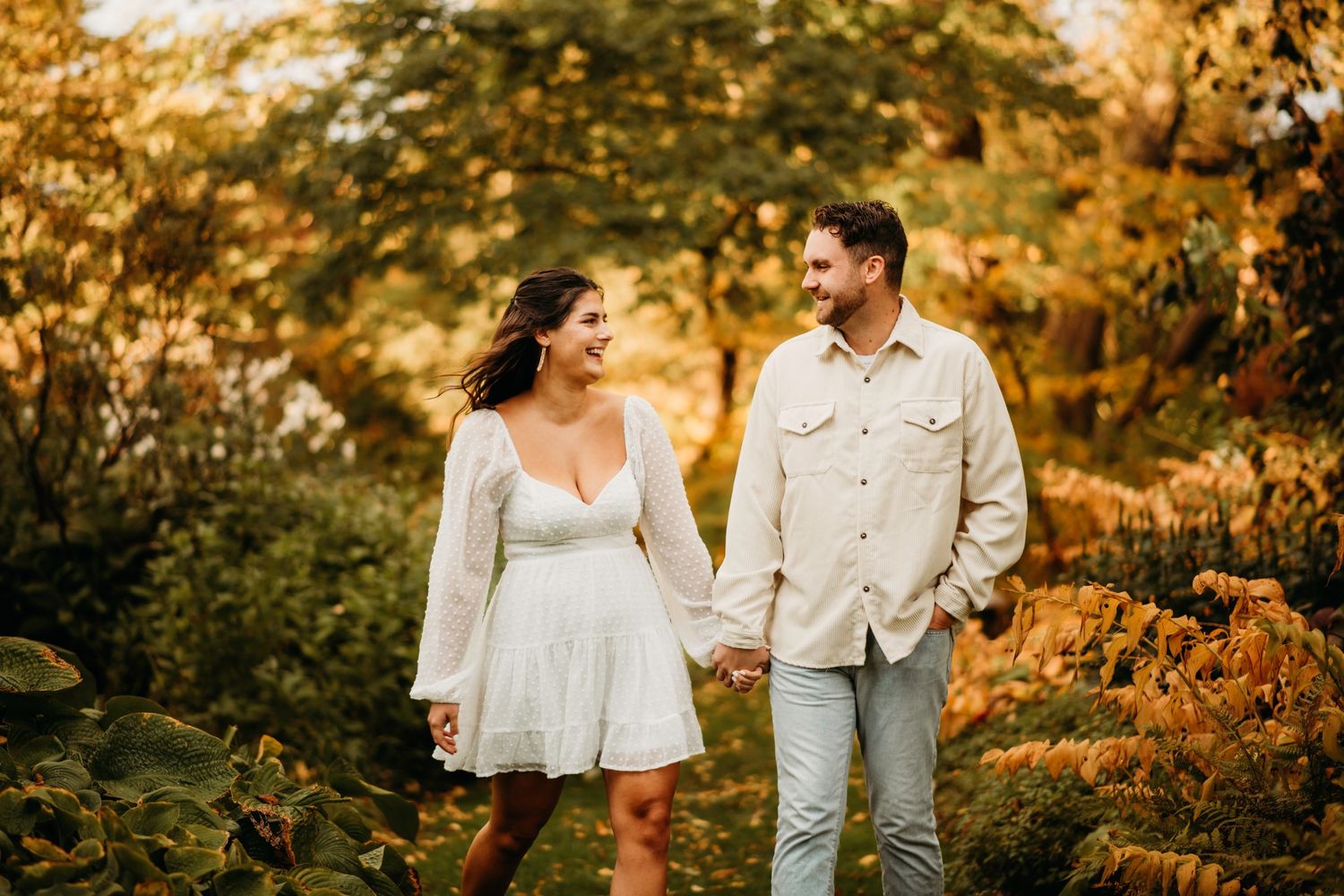 Couple holding hands and smiling while walking through autumn foliage in matching white outfits.