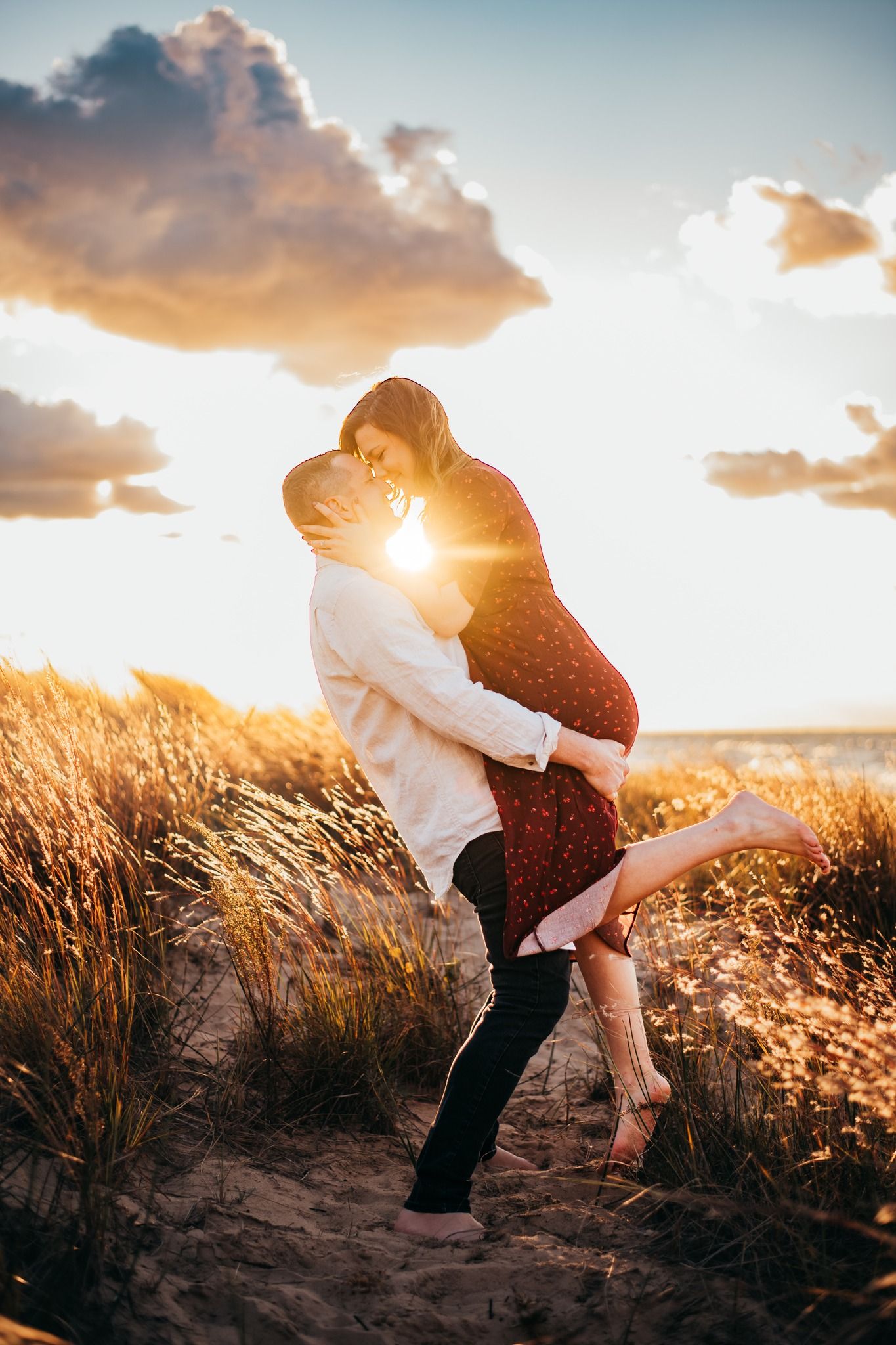 Man lifting woman at sunset on sandy beach dunes with dramatic clouds and golden light.