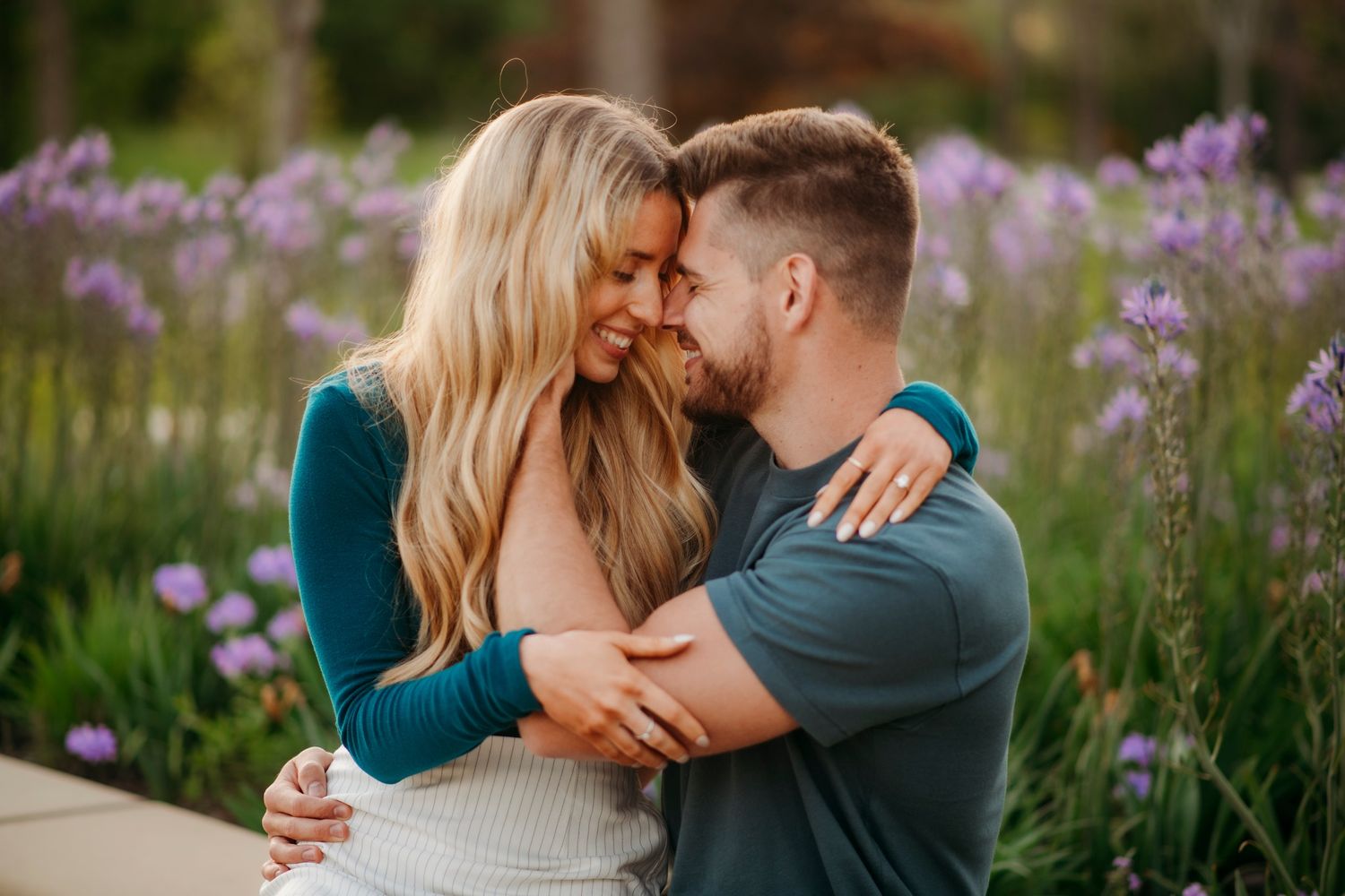 A couple embraces and touches foreheads romantically among purple wildflowers in a lush garden setting.
