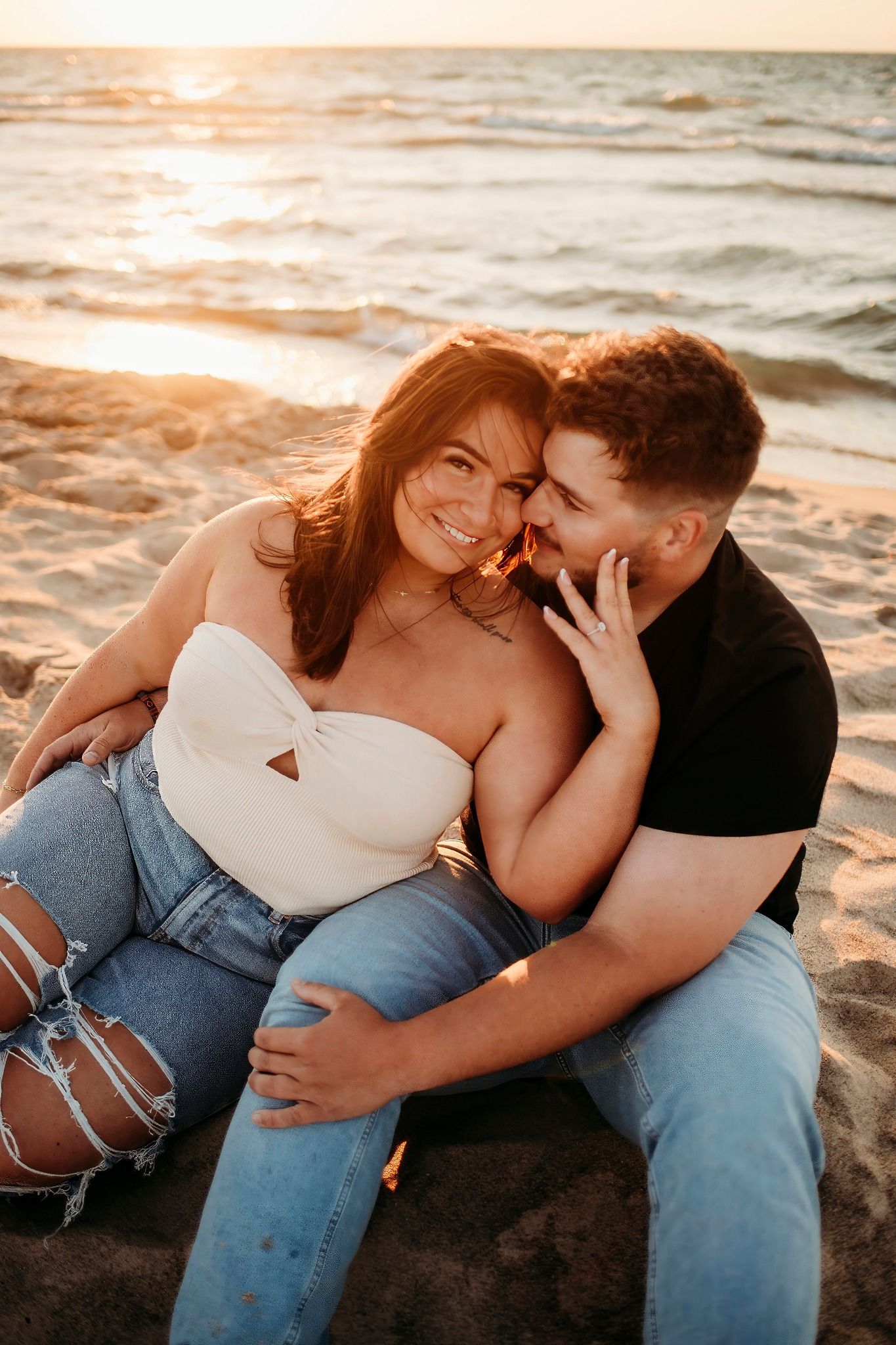 Couple sitting closely on beach at sunset, woman smiling while man whispers in her ear.