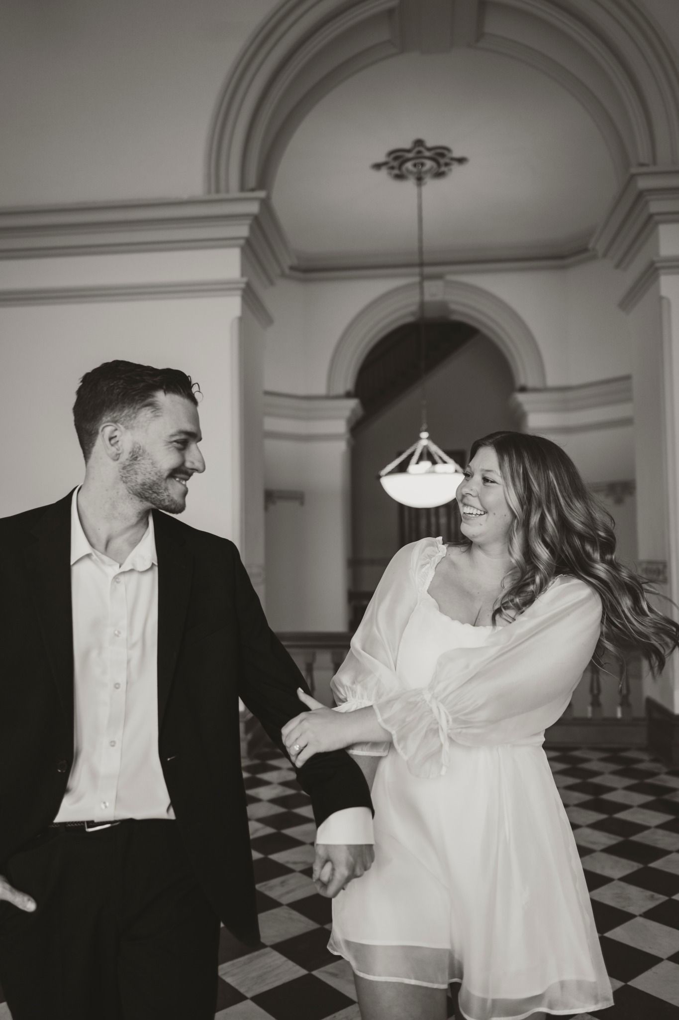 Black and white photo of a smiling couple holding hands in an elegant hall with arched ceilings and checkered floors.