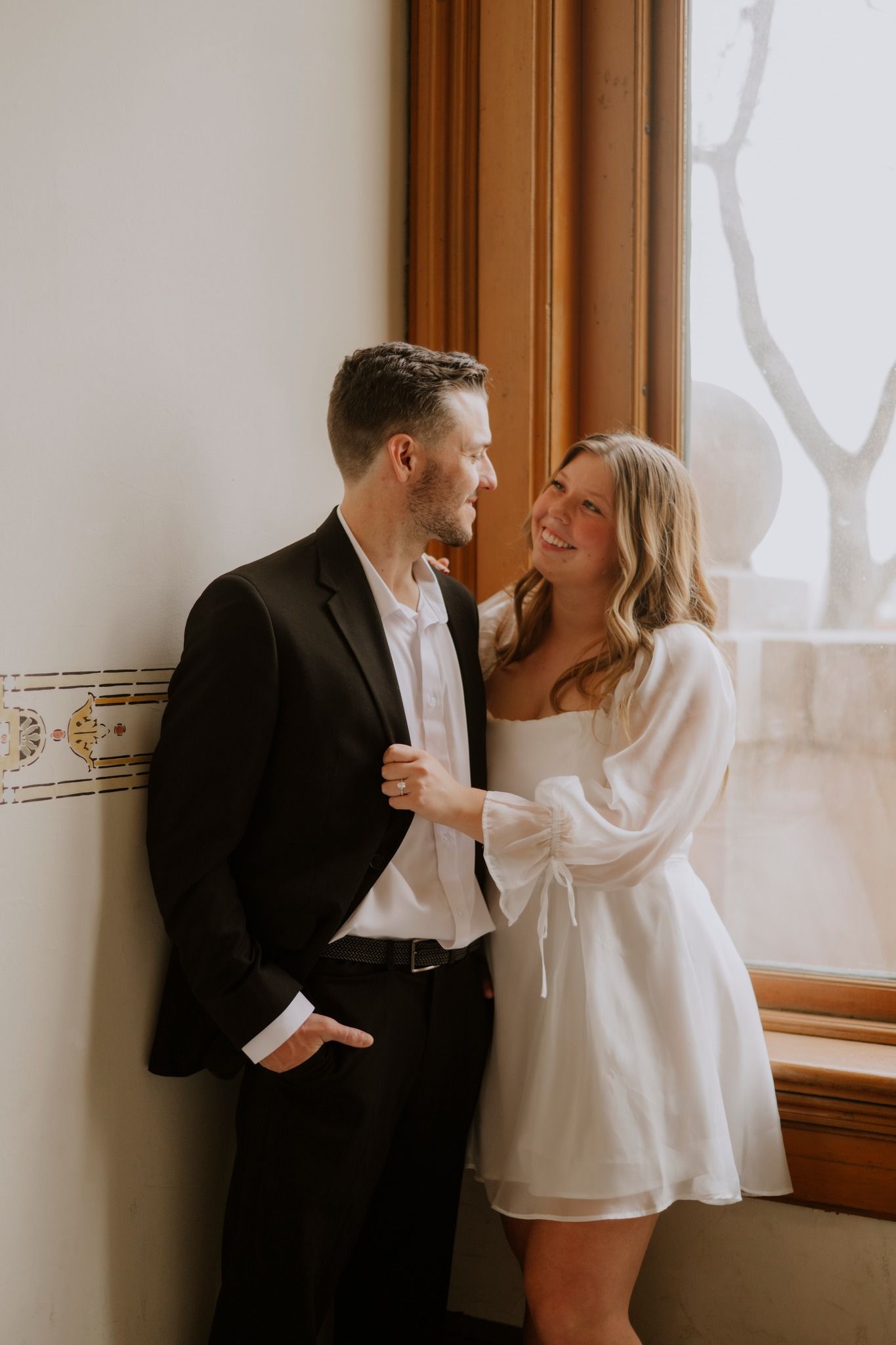 A happy couple in formal attire sharing a romantic moment by a sunlit window in a classic interior.