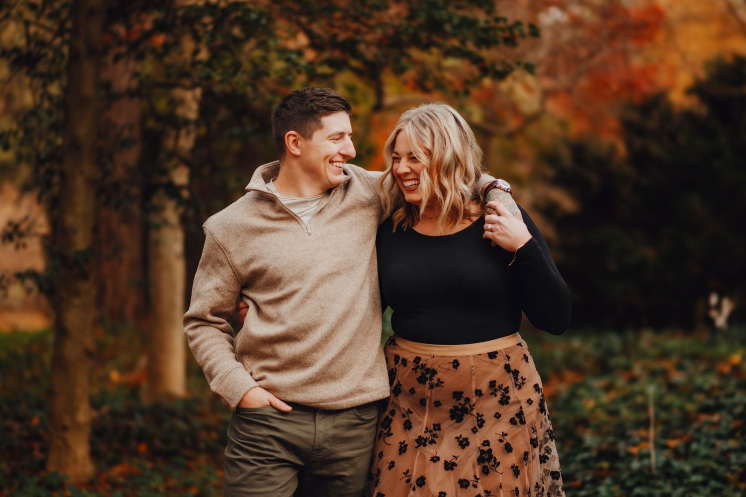 A couple laughs together outdoors in autumn, surrounded by colorful fall foliage during a romantic engagement photo session.