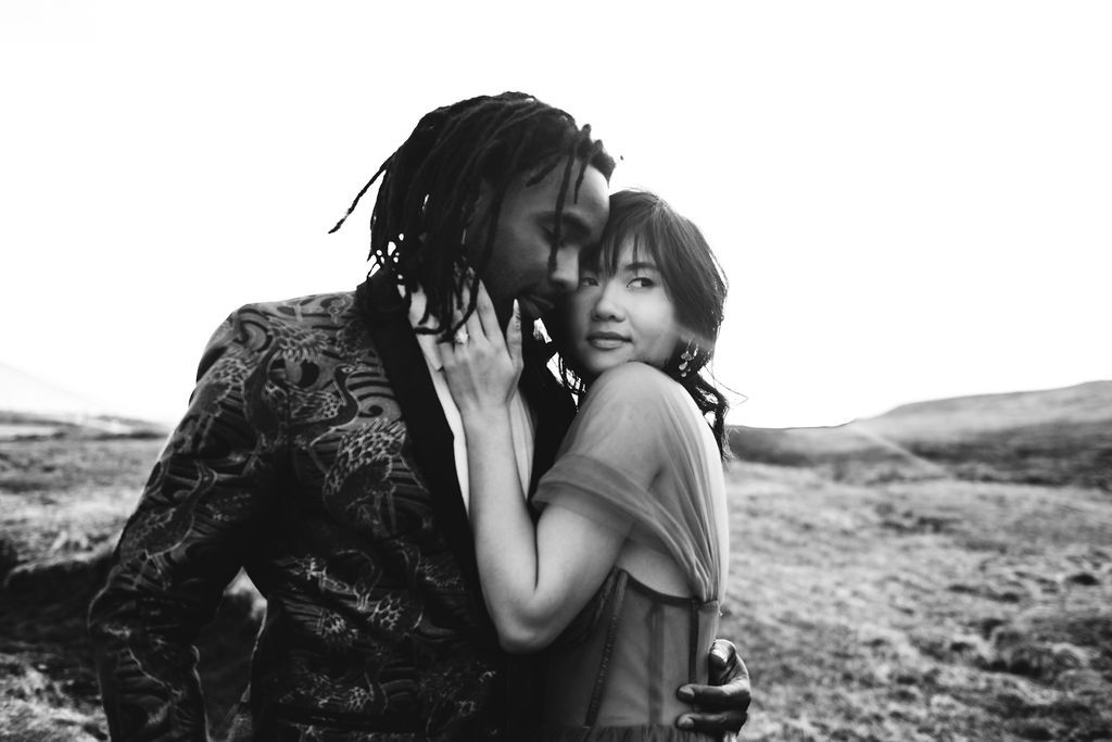 Black and white photo of a couple embracing tenderly outdoors in a scenic landscape.