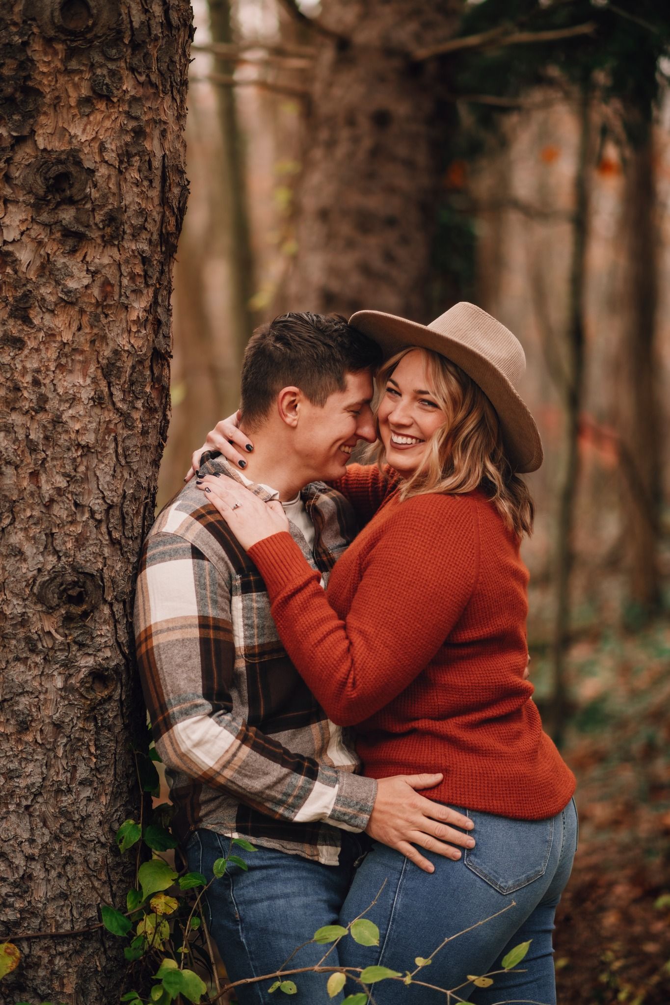 Couple embracing and laughing together in autumn forest, woman in rust sweater and hat, man in plaid shirt.