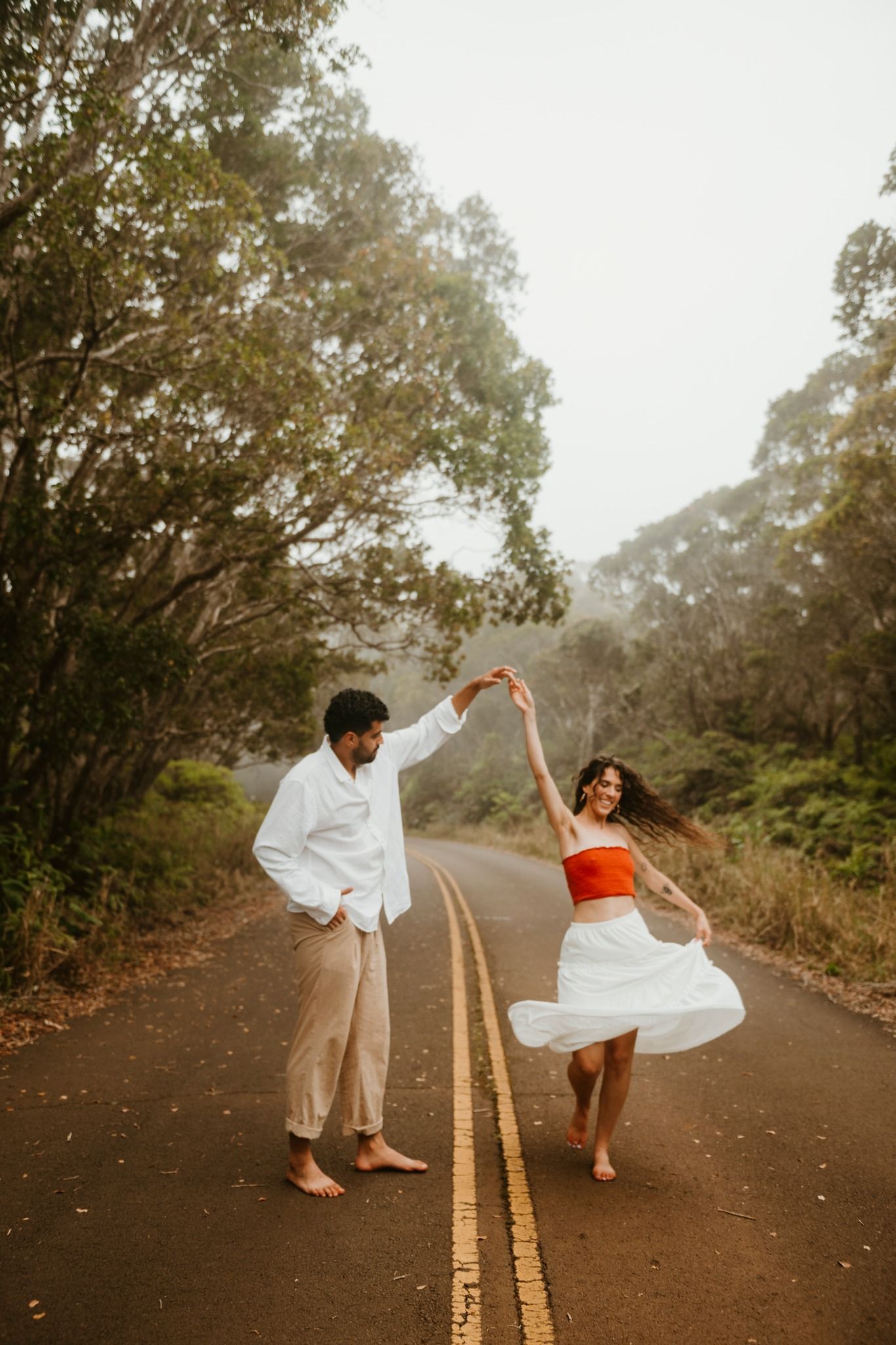 Couple dancing barefoot on a misty road surrounded by lush trees, woman in red top and white skirt, man in white shirt.